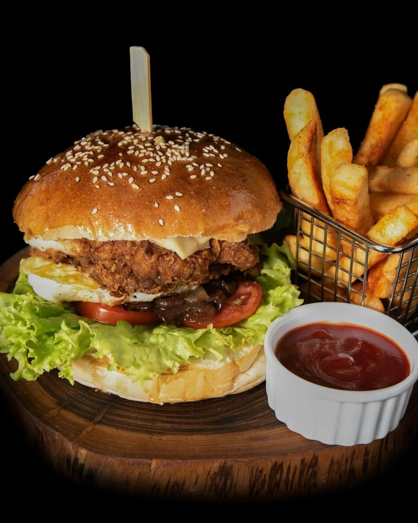 A close-up of a fried chicken sandwich with lettuce, tomato, and mayonnaise, served with French fries, a small container of ketchup, and a cup of ketchup on a wooden board.