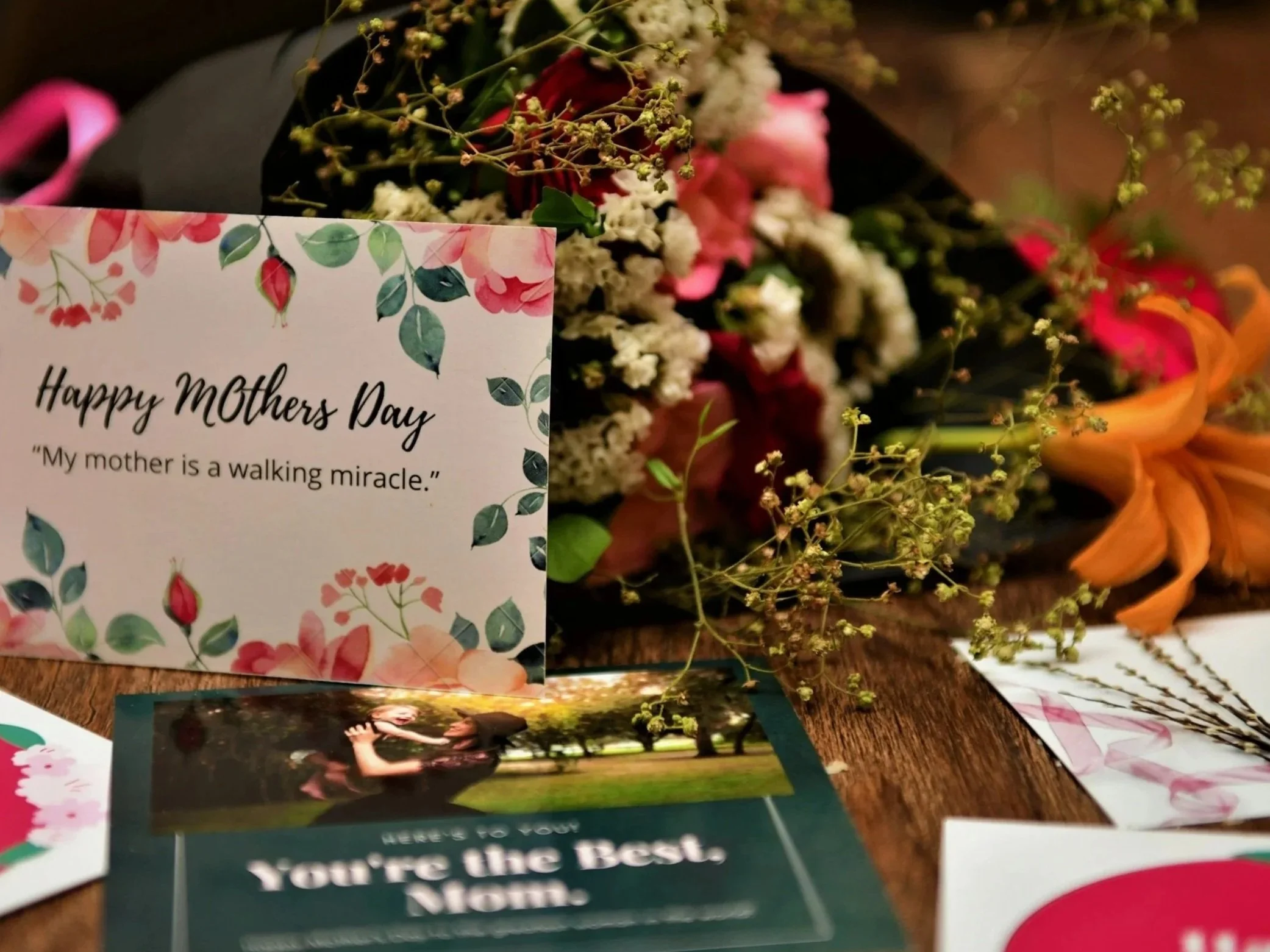 A bouquet of flowers with white, pink, and orange blooms surrounds a graduation card that reads "Happy Mother's Day." The card features a floral design with green leaves and pink flowers. A photograph of a girl and woman hugging outdoors is also visible on the table.