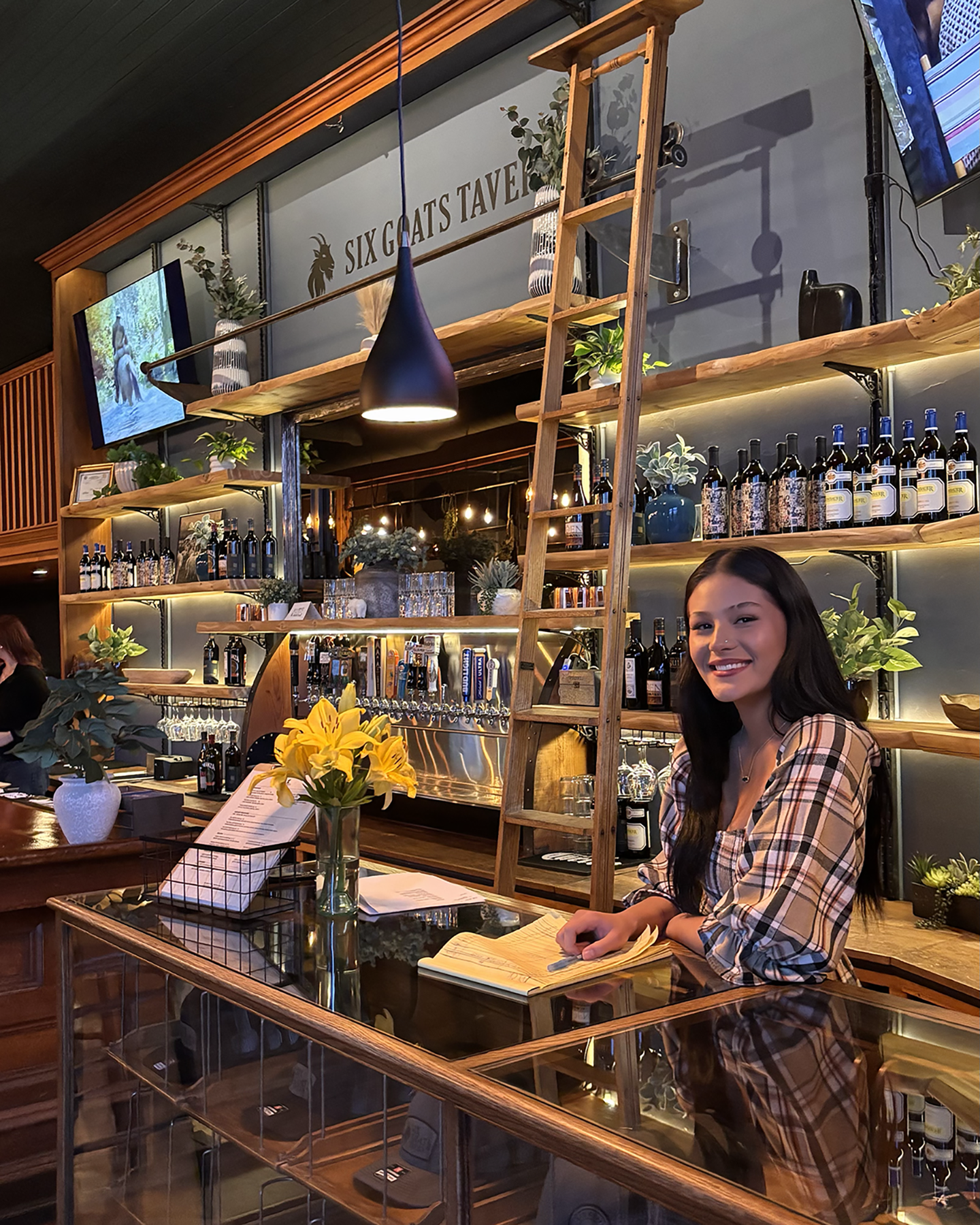 A woman with long dark hair wearing a plaid shirt, smiling while greeting guests at Six Goats Tavern in Spearfish, South Dakota. The bar is decorated with plants, wine bottles, and a ladder, with a TV mounted on the wall showing sports.