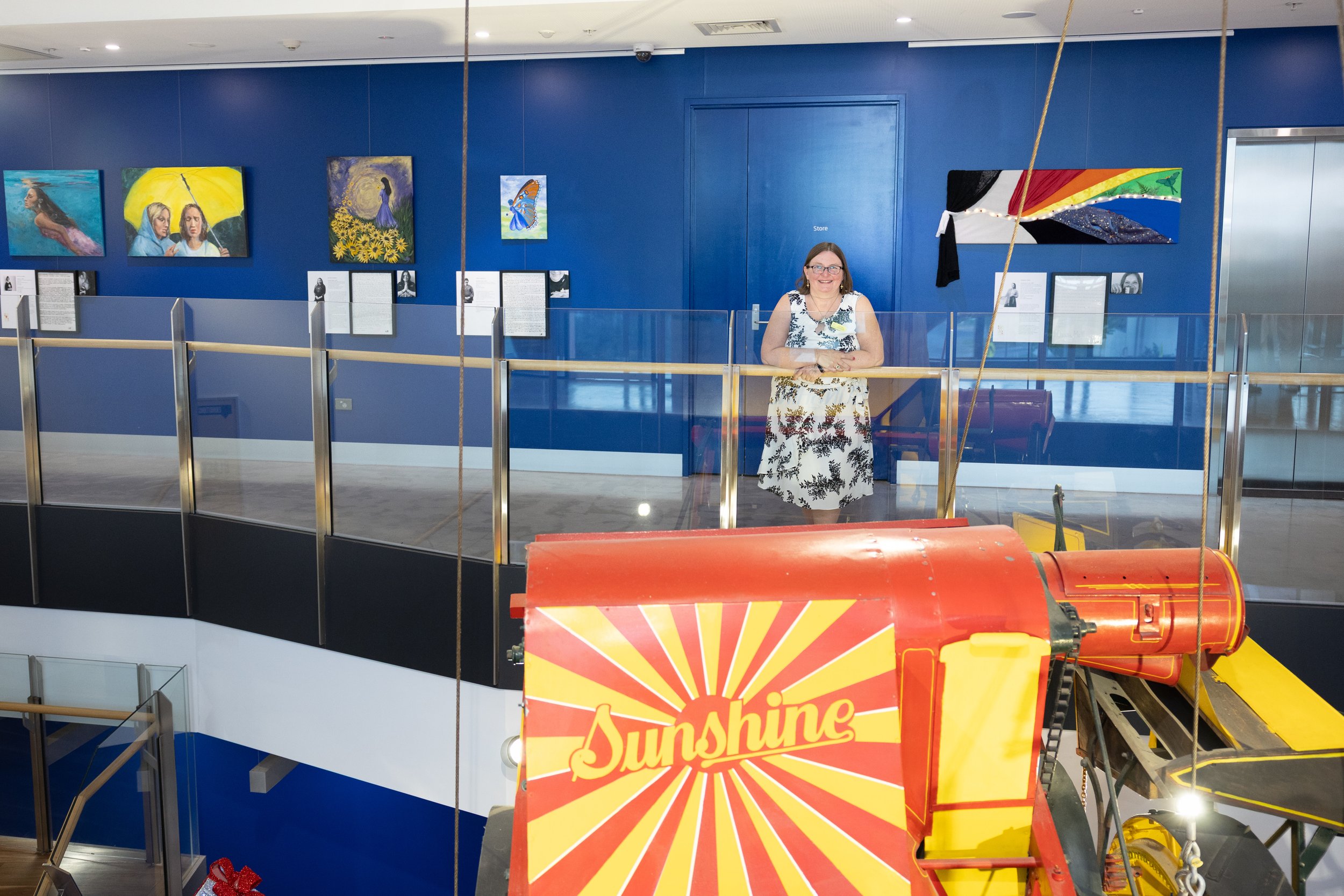 A woman standing behind a glass barrier in an art gallery, with paintings on a blue wall behind her and a colorful kite with a rainbow pattern on a display structure in the foreground.