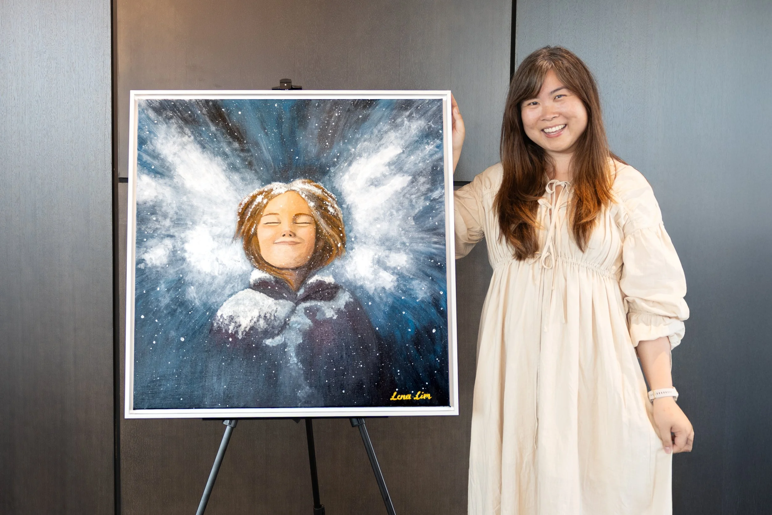 A woman standing next to a large painting of a girl with closed eyes, smiling, and surrounded by a cosmic, starry background. The painting is signed by Lena Lim.