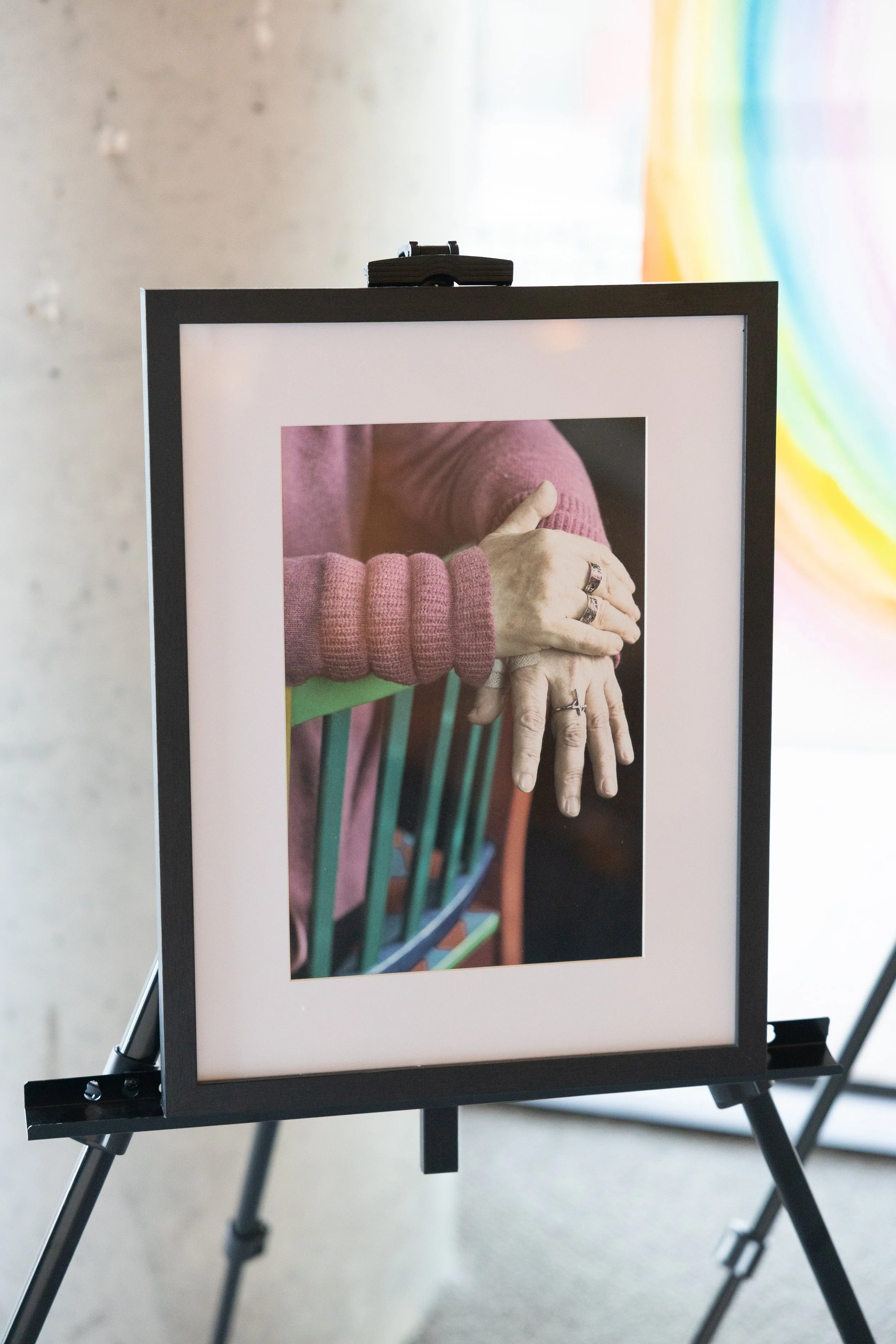 Photograph of an art display, showing a close-up of a person's hands resting on the back of a colorful chair, with visible rings on their fingers. The person is wearing a pink sweater, and the background includes a blurred rainbow-colored mural.