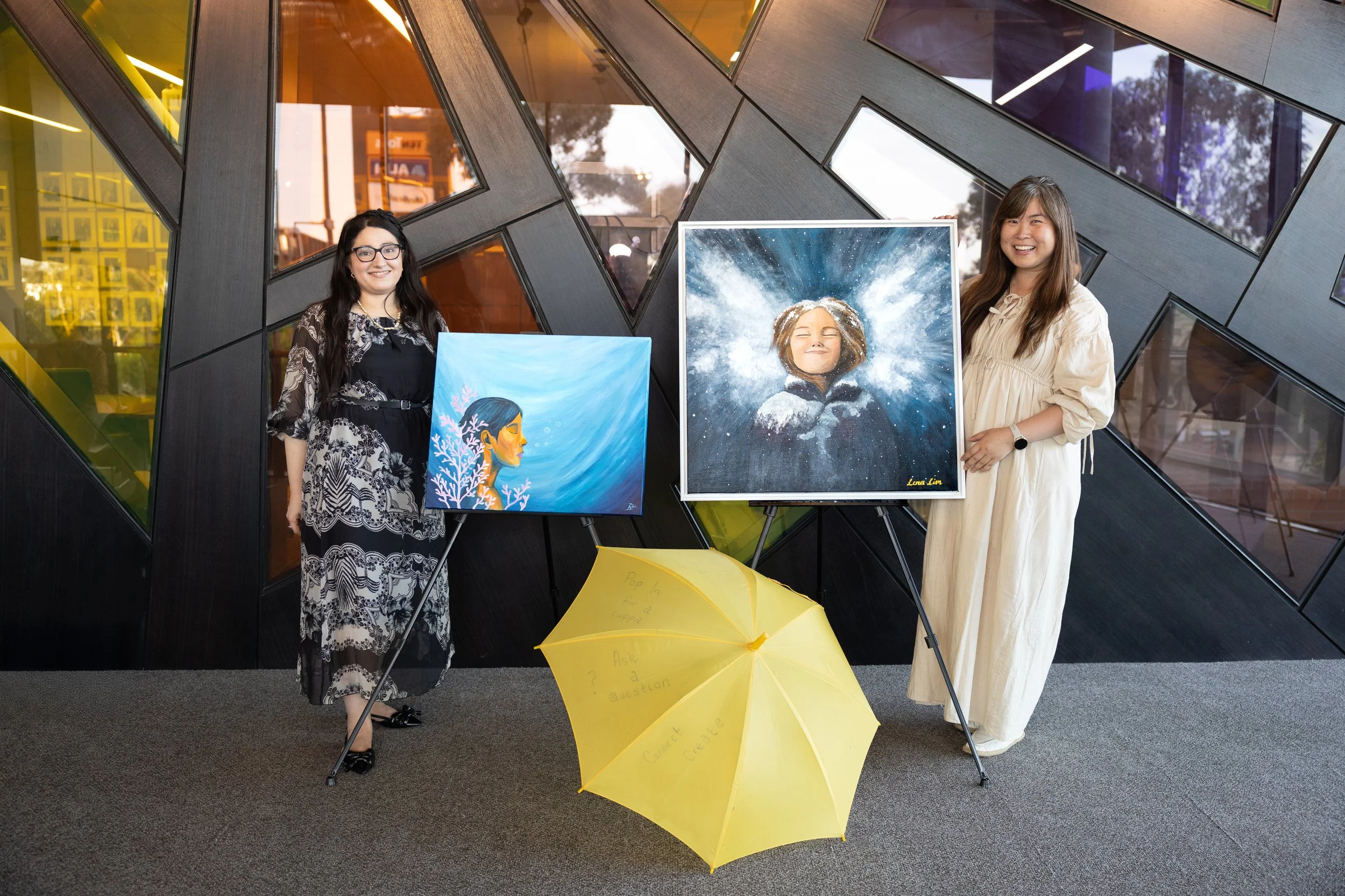 Two women stand next to display easels with two paintings, in front of a geometric black wall with glass panels. A yellow umbrella is on the floor in front of them.