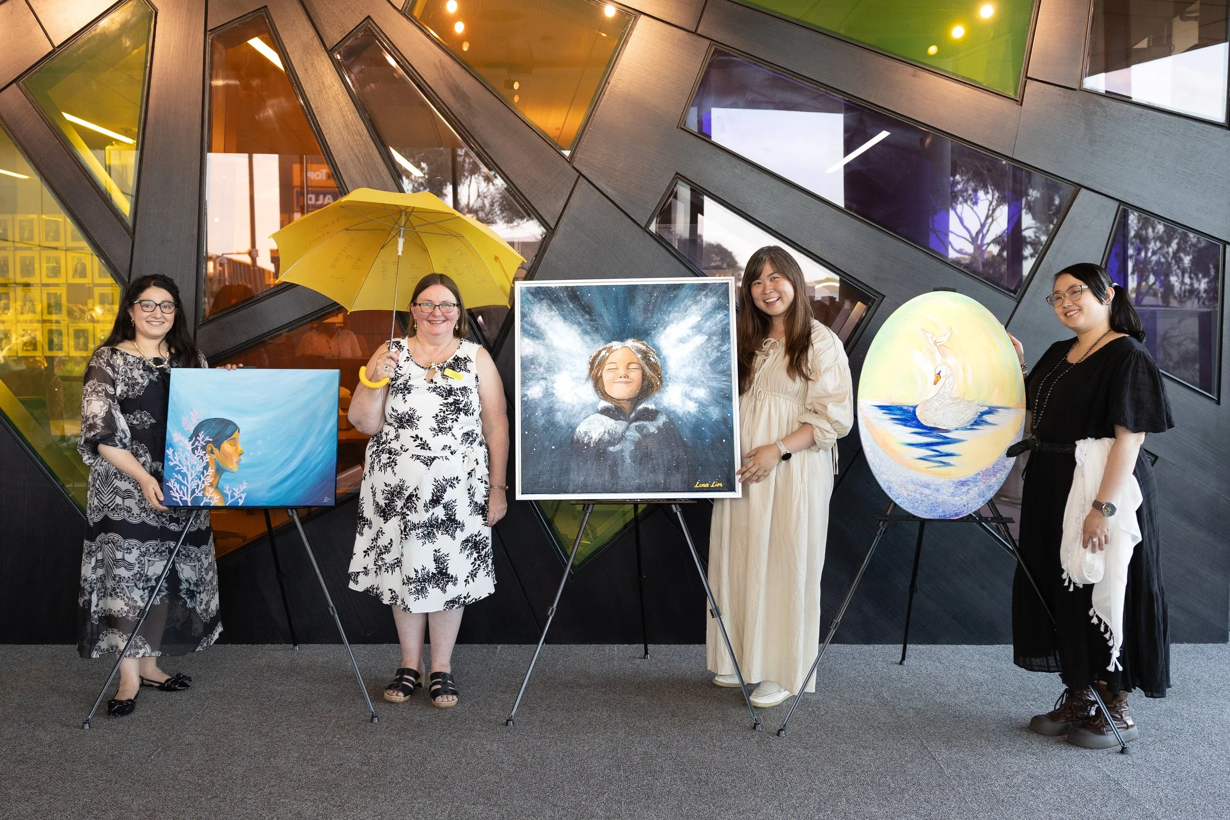 Four women stand indoors in front of a modern, colorful, geometric wall, each holding or standing by a piece of artwork featuring swan motifs or images of water. The second woman from the left holds a yellow umbrella. The artwork showcases a woman wi