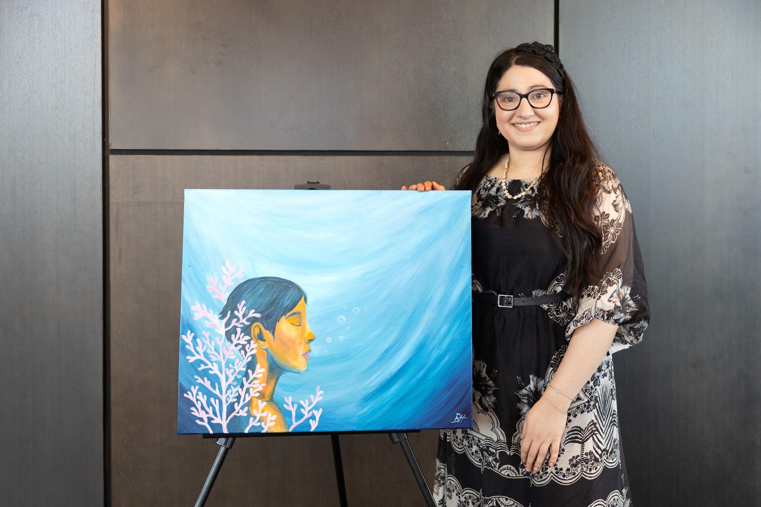 A woman with glasses and long dark hair holding a painting on an easel. The painting depicts a profile of a woman with short dark hair underwater, with pink coral branches around her. The background is blue with wave-like strokes.
