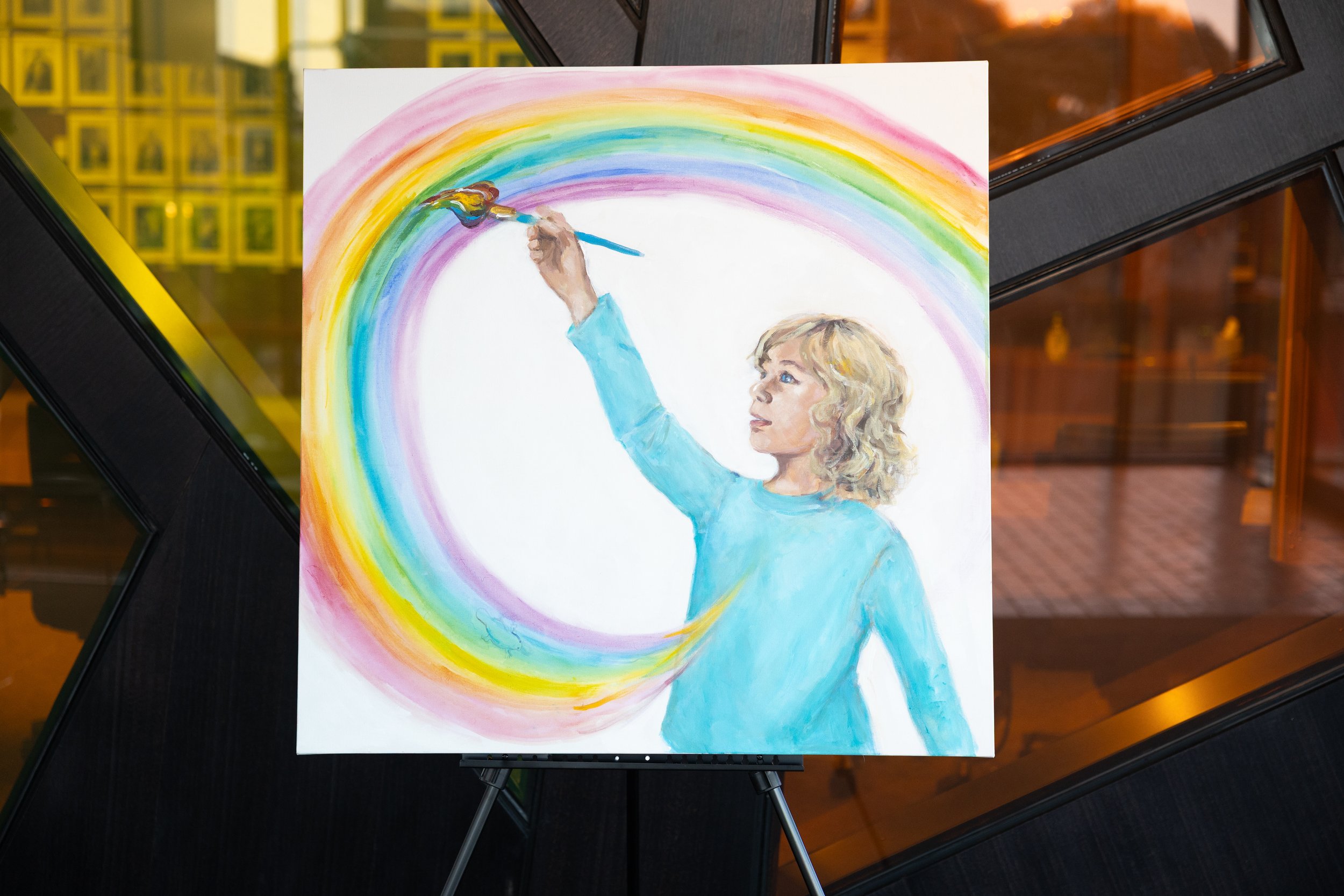 Painting of a woman with blonde curly hair in a blue shirt, raising a paintbrush with colorful paint to create a rainbow on a white canvas.