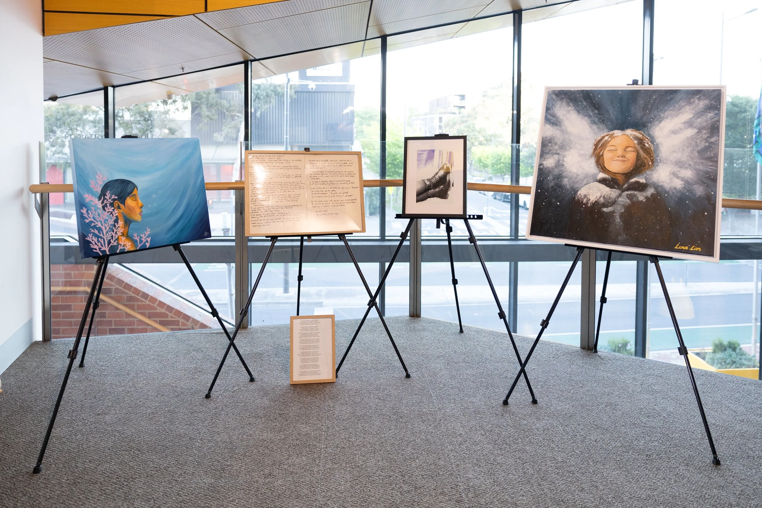 An art gallery display featuring four paintings on easels, with a report or description on a stand in front of them, inside a modern building with large windows and cityscape view.