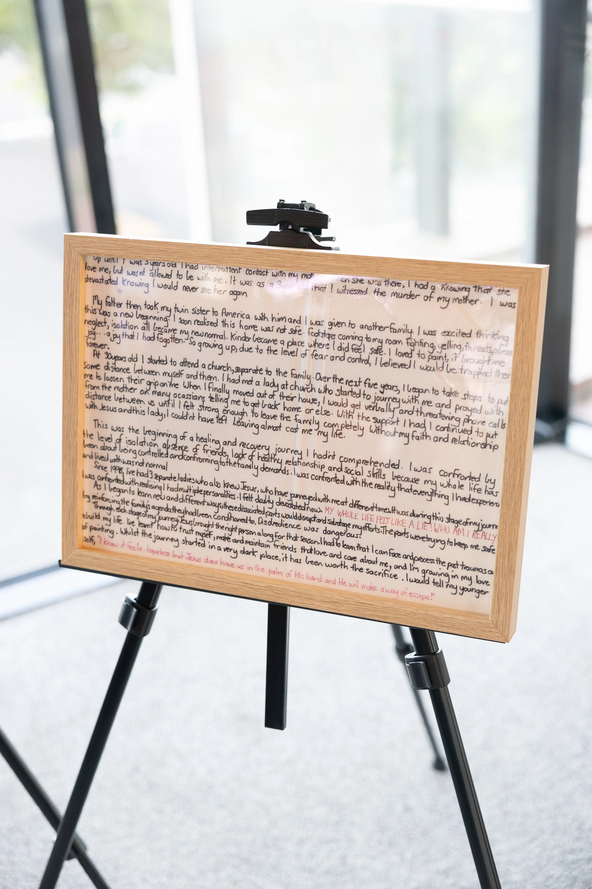 A framed handwritten letter on a wooden easel, with visible text in black and red ink, displayed in a room with large windows in the background.