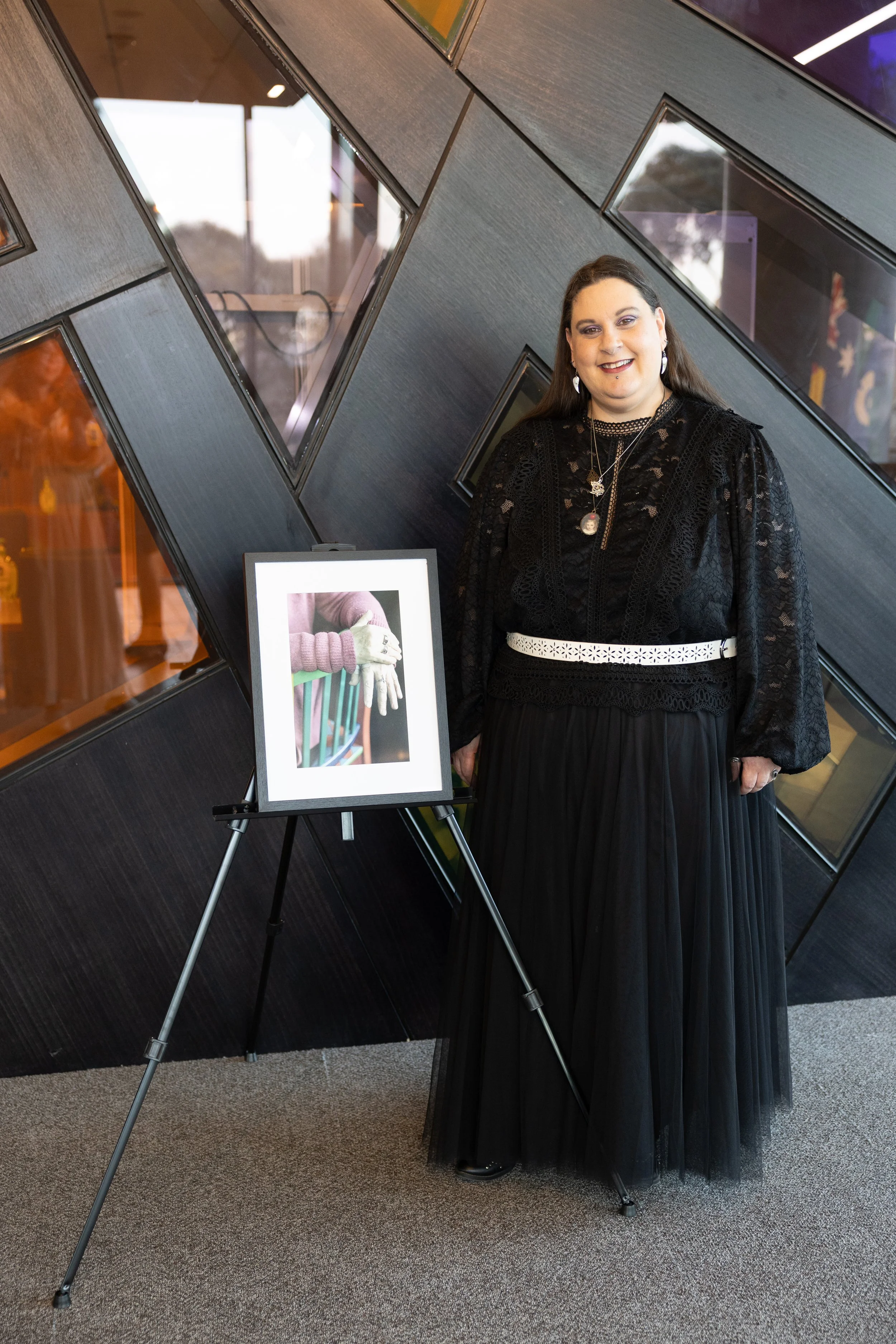 A woman dressed in a black lace top and a black pleated skirt, standing next to a framed picture of a pair of hands wearing white gloves, holding a small object while resting on a railing, at an indoor art exhibit or gallery with geometric black wall