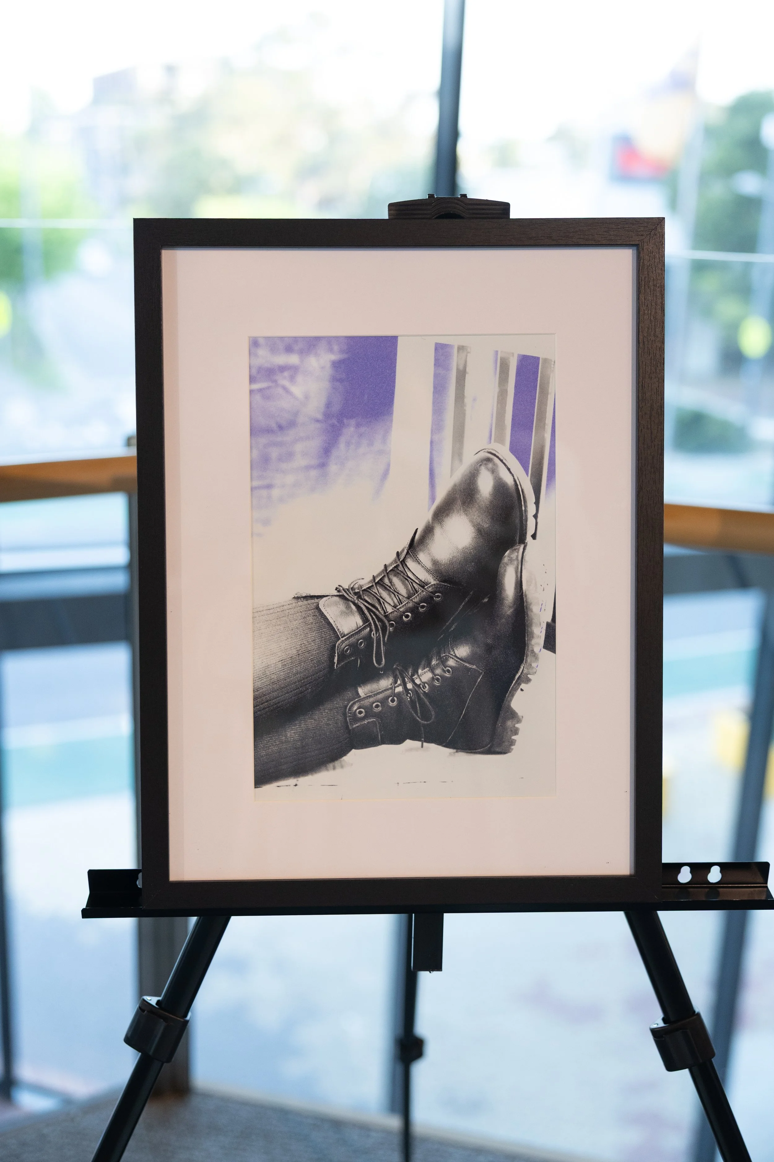 A framed black and white photograph of person wearing hiking boots, lying on their back with legs elevated against a railing, displayed on an easel indoors with windows and a blurred outdoor scene in the background.