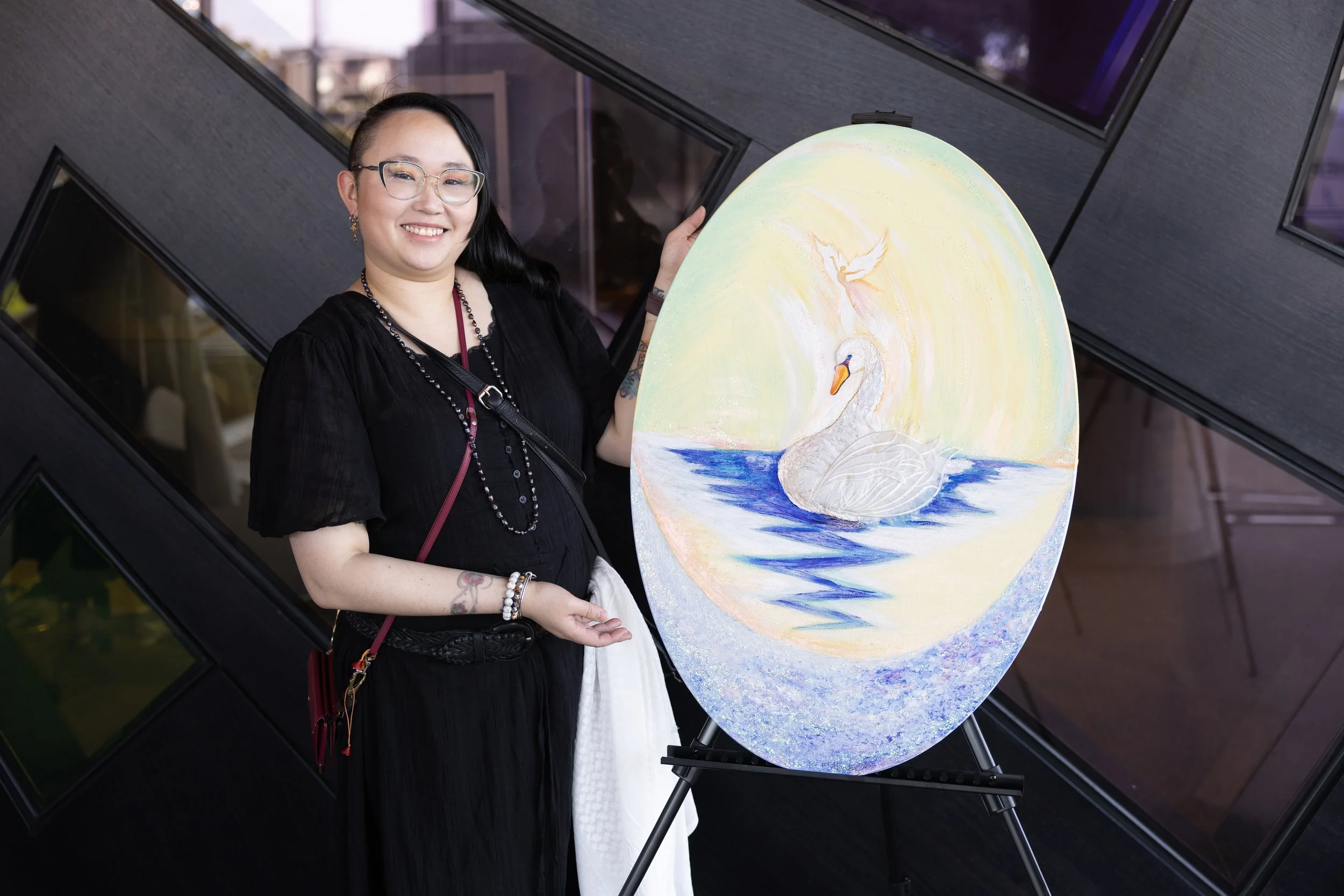 A woman with black hair, glasses, and a black dress smiling while holding a swan painting on a large oval canvas.