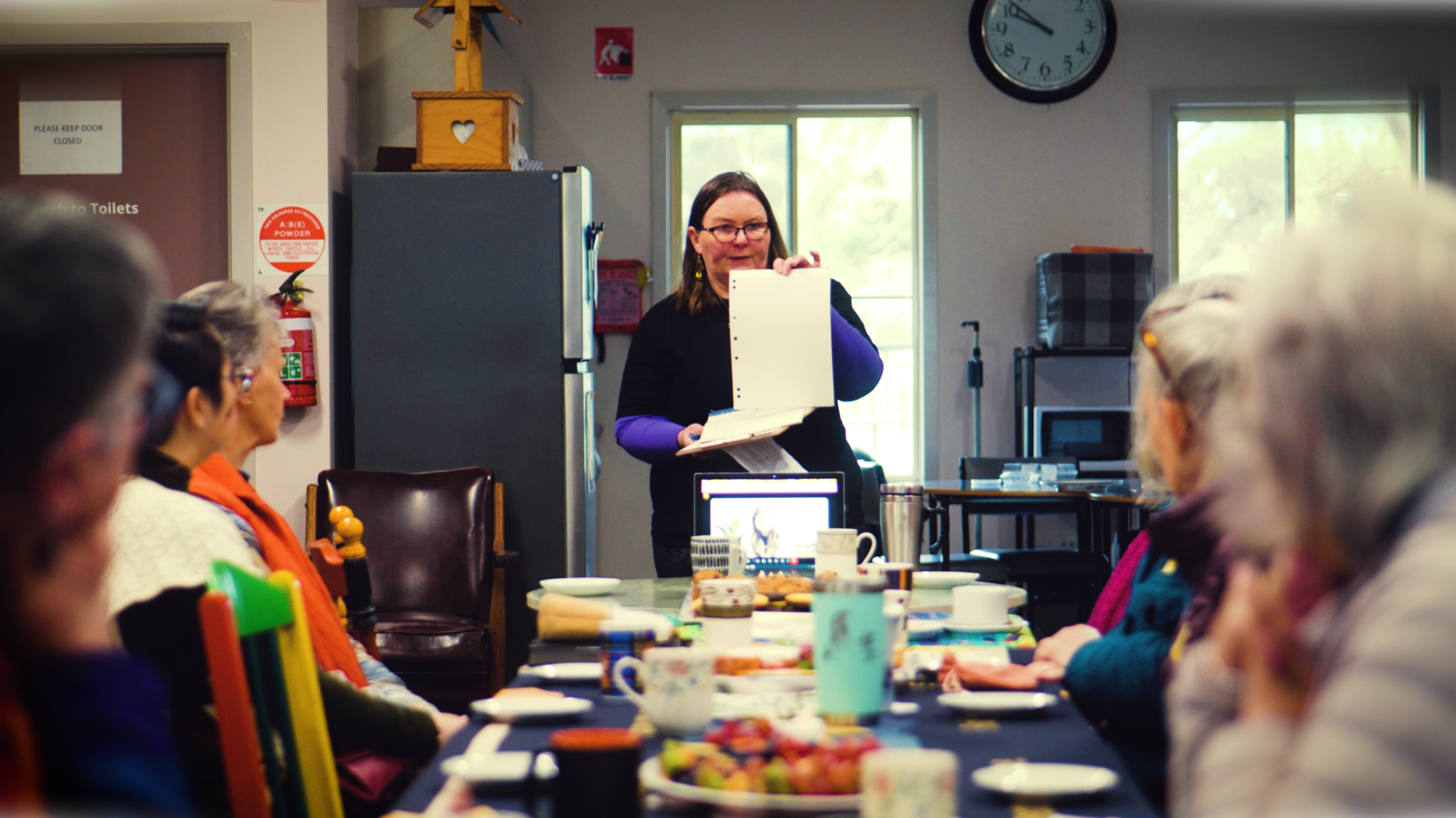 A woman standing at the head of a table, holding an open binder, leading a group of elderly women in a meeting or discussion in a bright room with windows, clock, and kitchen appliances.