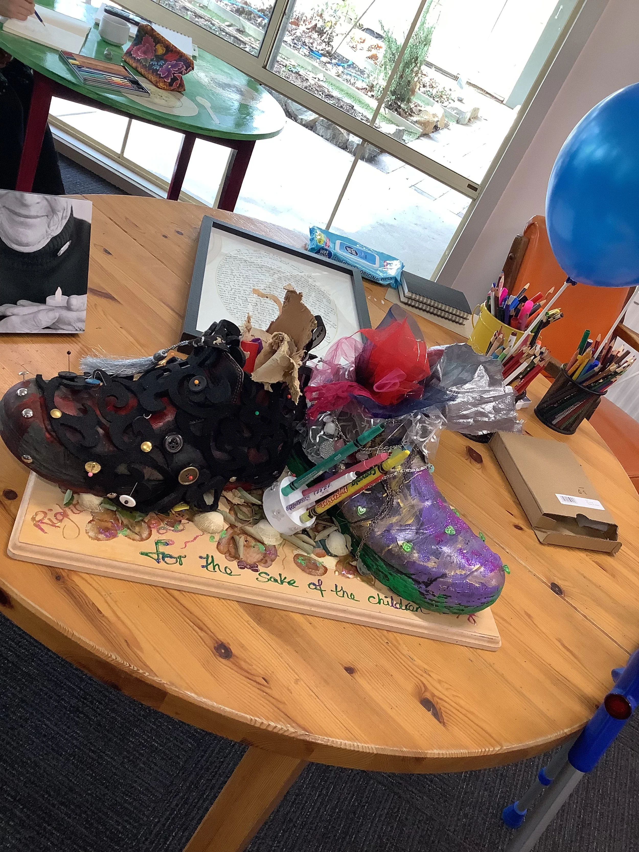 A decorated cake shaped like a shoe on a wooden table, surrounded by markers, pens, and craft supplies, with a framed photo, balloons, and handwritten message.