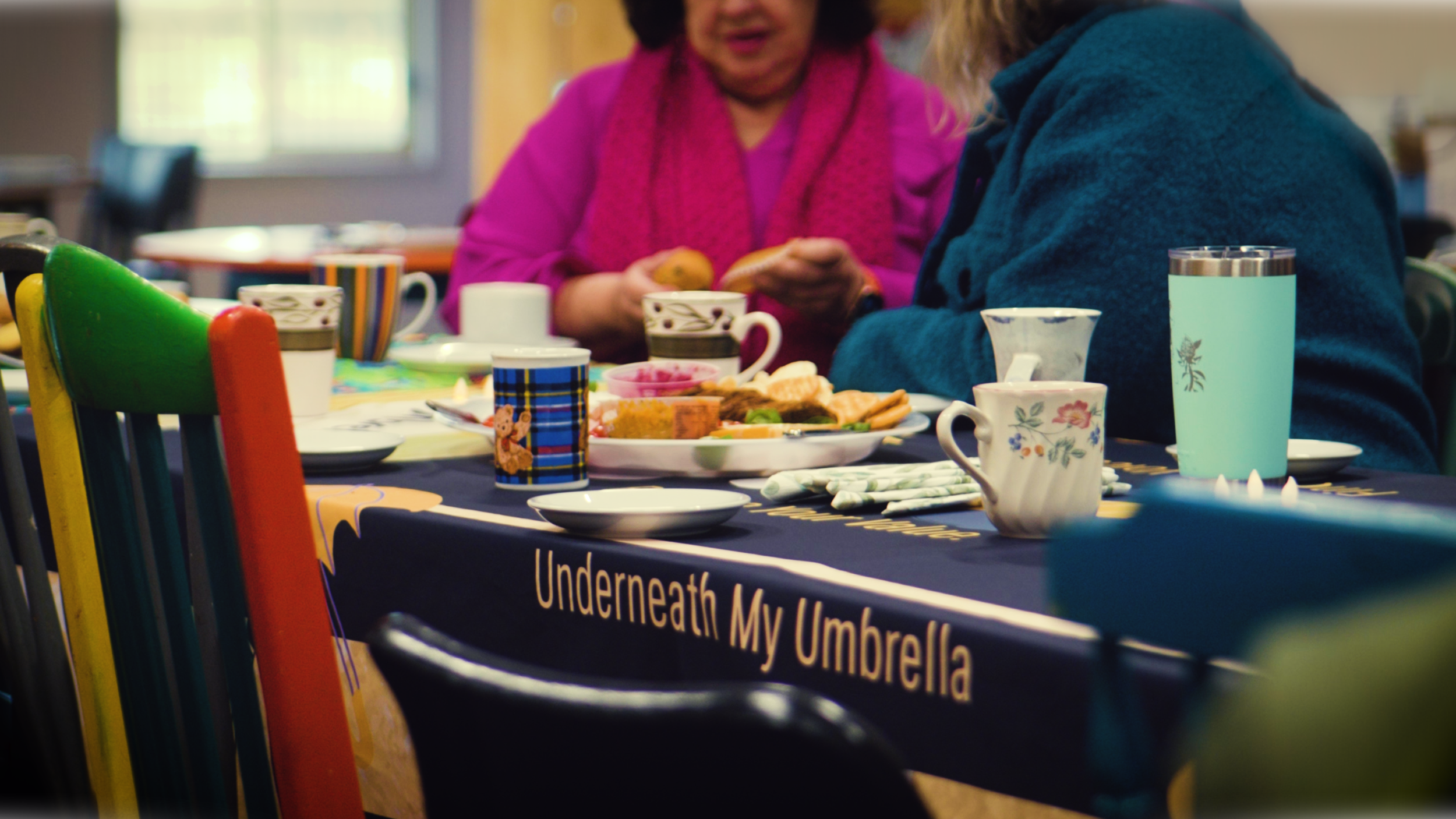 Two women sitting at a table with breakfast foods, cups, and mugs, in a cheerful setting with colorful chairs and a black tablecloth that reads 'Underneath My Umbrella.'