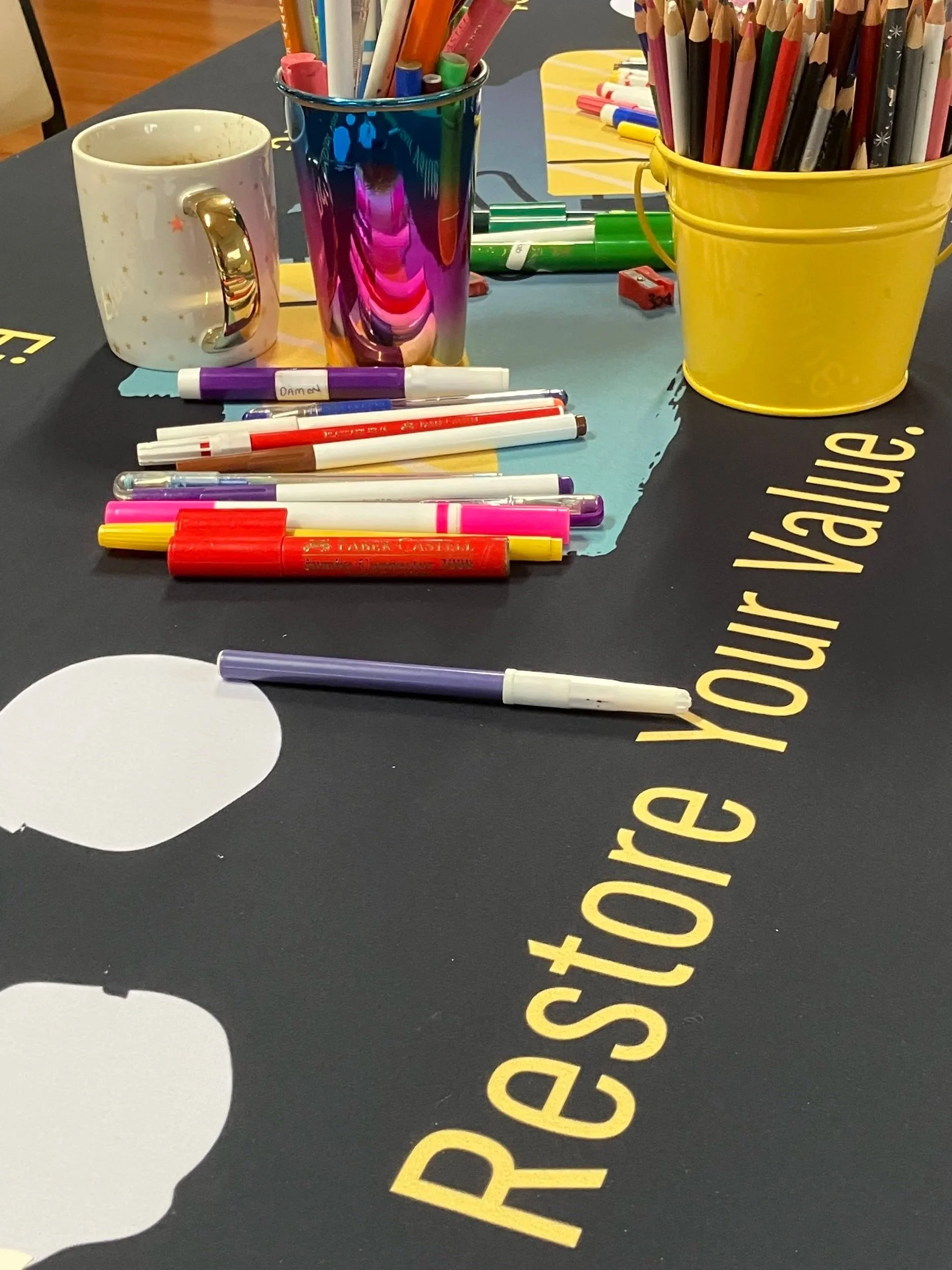 A table with colorful markers, pens, and pencils in cups, a white mug, and a yellow bucket, with the words 'Respect your value' on a black surface.