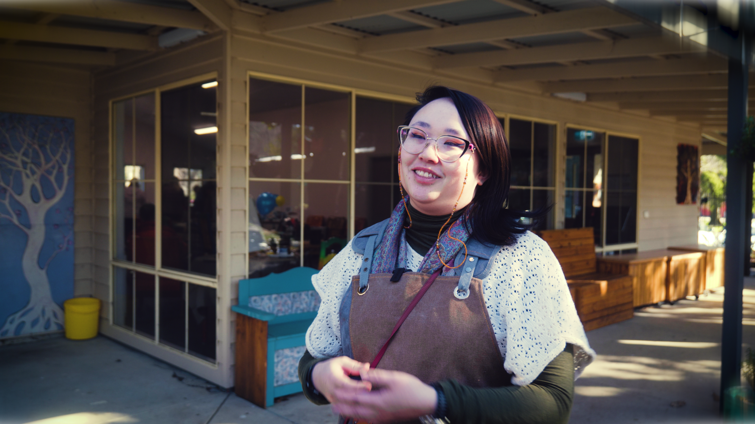A woman with glasses and earrings standing outdoors in front of a building with large windows, smiling and holding her hands together, wearing a brown apron and scarf.