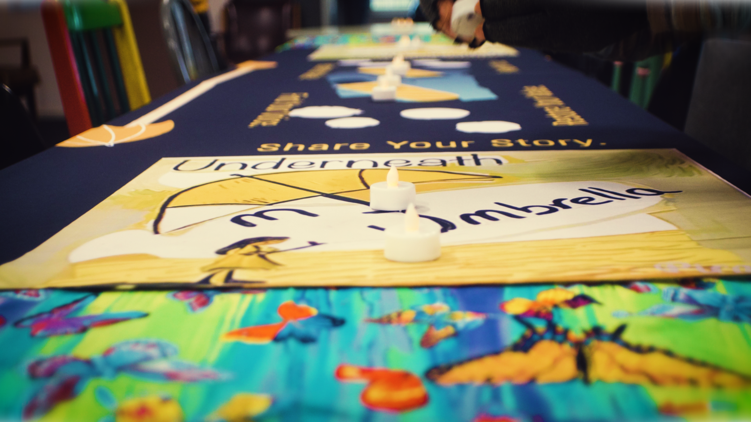 A colorful table cover featuring butterflies and flowers, with candles and a banner that says 'Share Your Story' and 'Underrneath' visible. The focus is on the candles and part of the banner, with chairs and blurred background in a room.