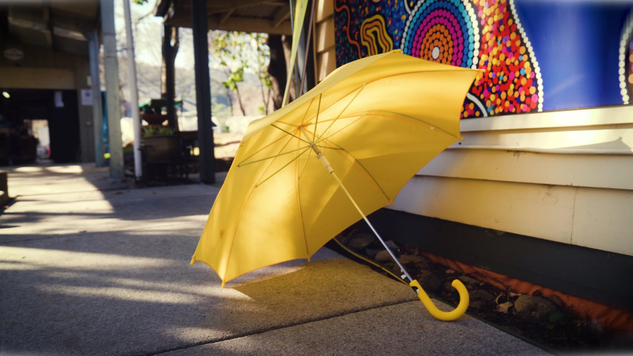 Yellow umbrella on a concrete sidewalk, leaning against a bench or wall with colorful artwork in the background.