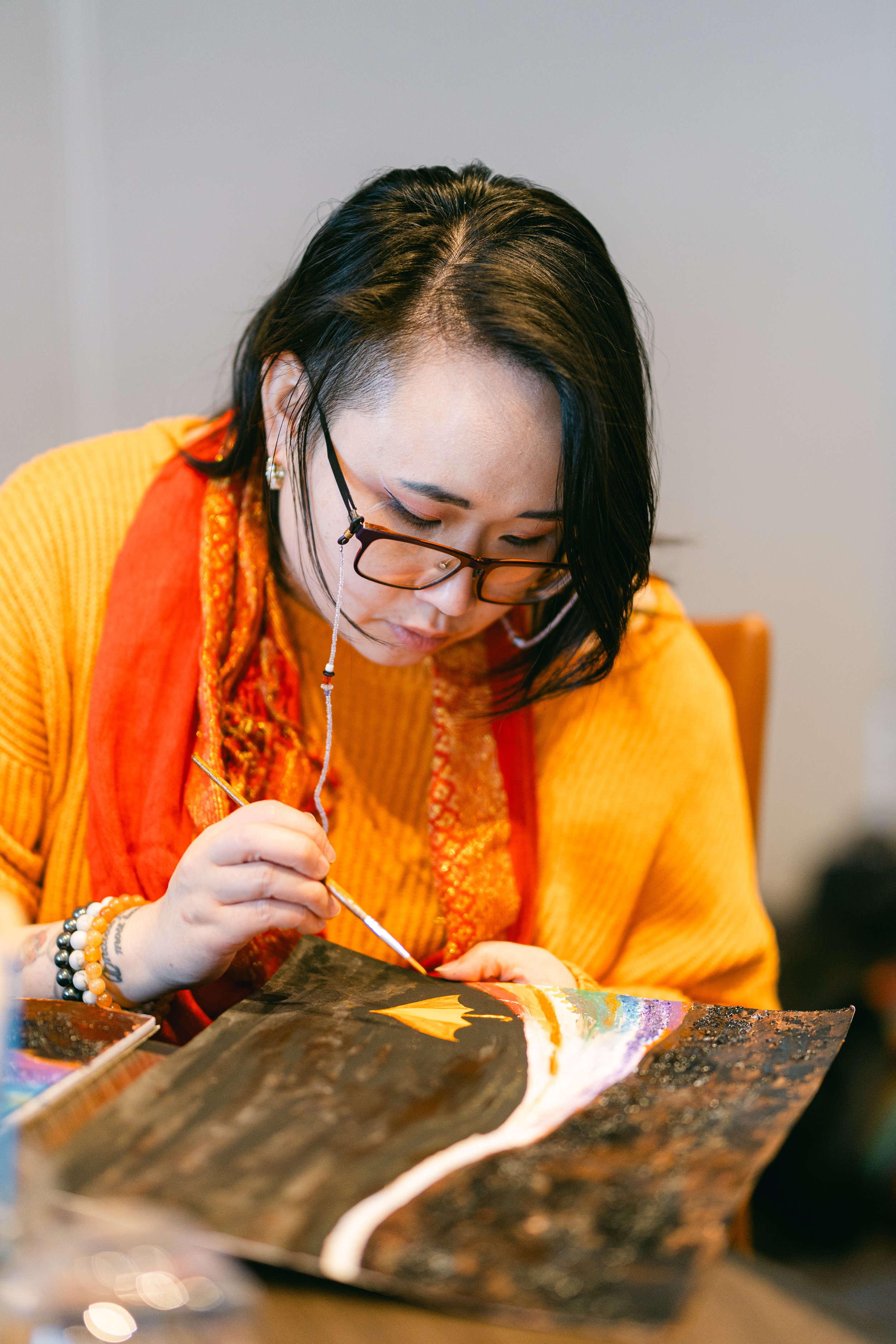 Woman in glasses with black hair, painting on a canvas with bright colors, wearing an orange sweater and a red scarf.