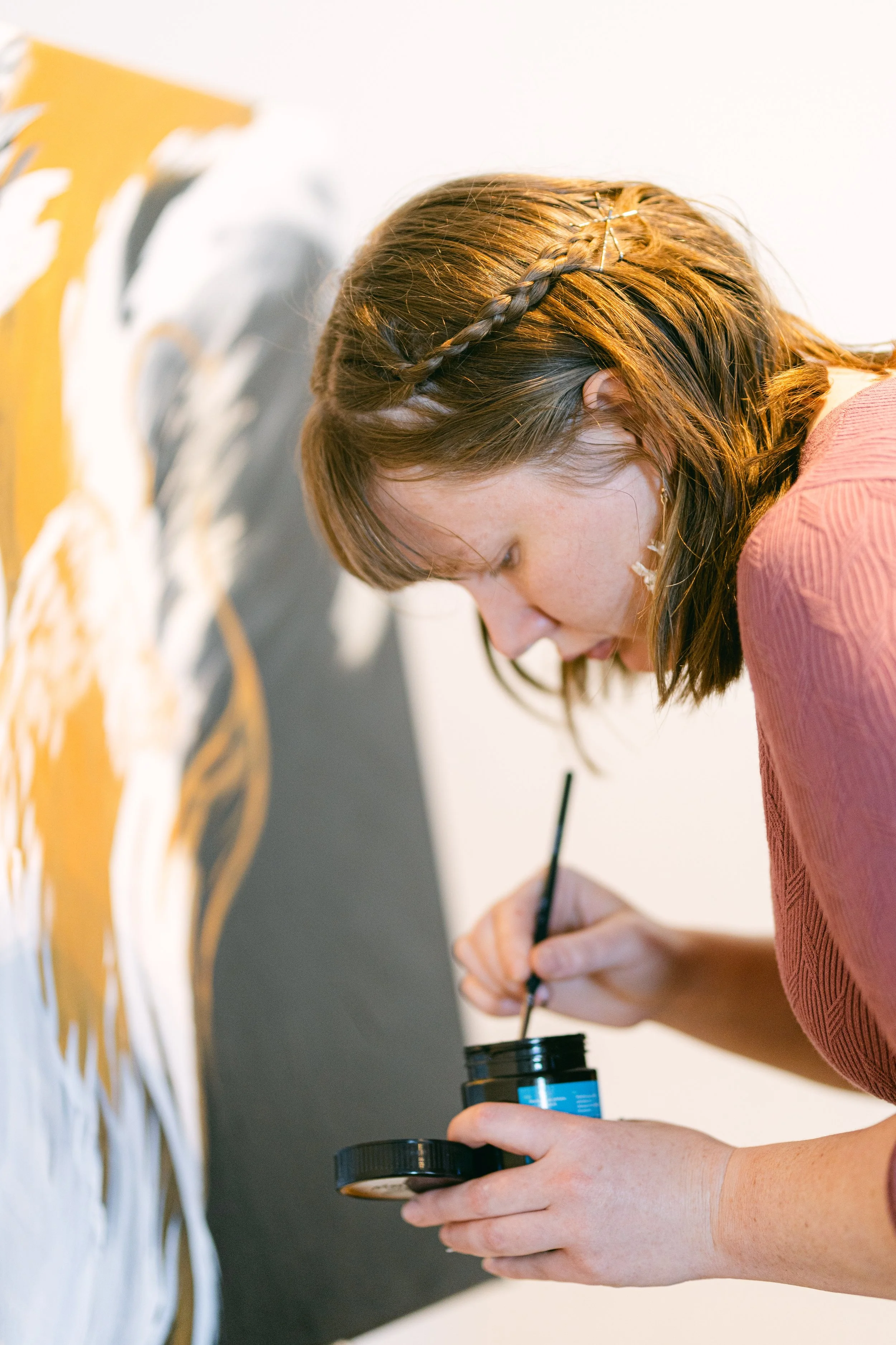 Young woman with brown hair, wearing a reddish top, painting a mural or artwork on a wall with black and white colors, holding a small jar of paint and a paintbrush.