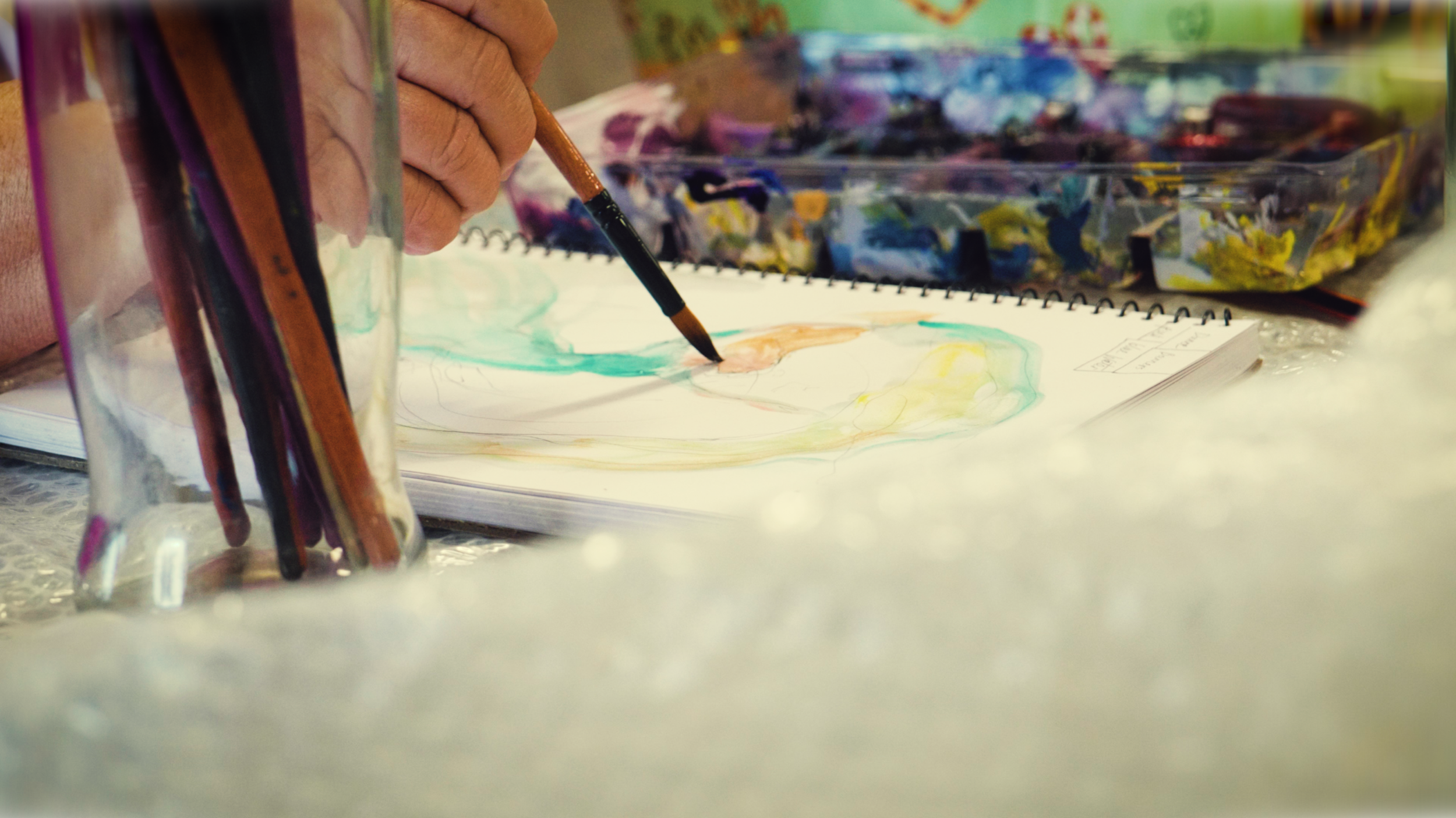 Hand holding a paintbrush, watercolor painting on sketchbook, art supplies in the background.