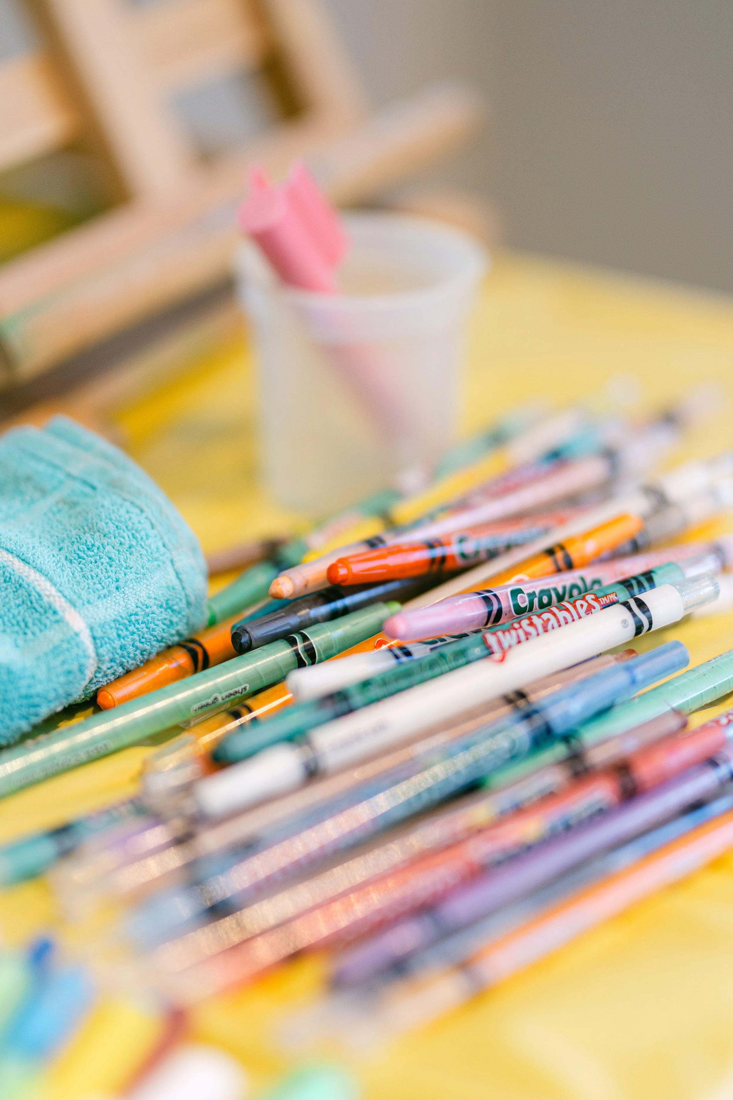 A collection of colorful markers and pens spread out on a yellow surface, with a folded towel and a white container holding pink objects in the background.