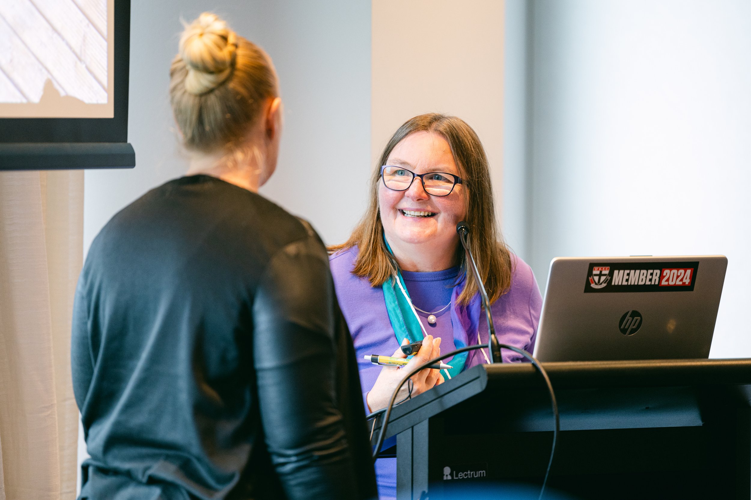 A woman with glasses and a purple shirt smiling at a woman with her back to the camera, at a conference or seminar, with a laptop that has stickers reading 'MEMBER 2024' and the HP logo.