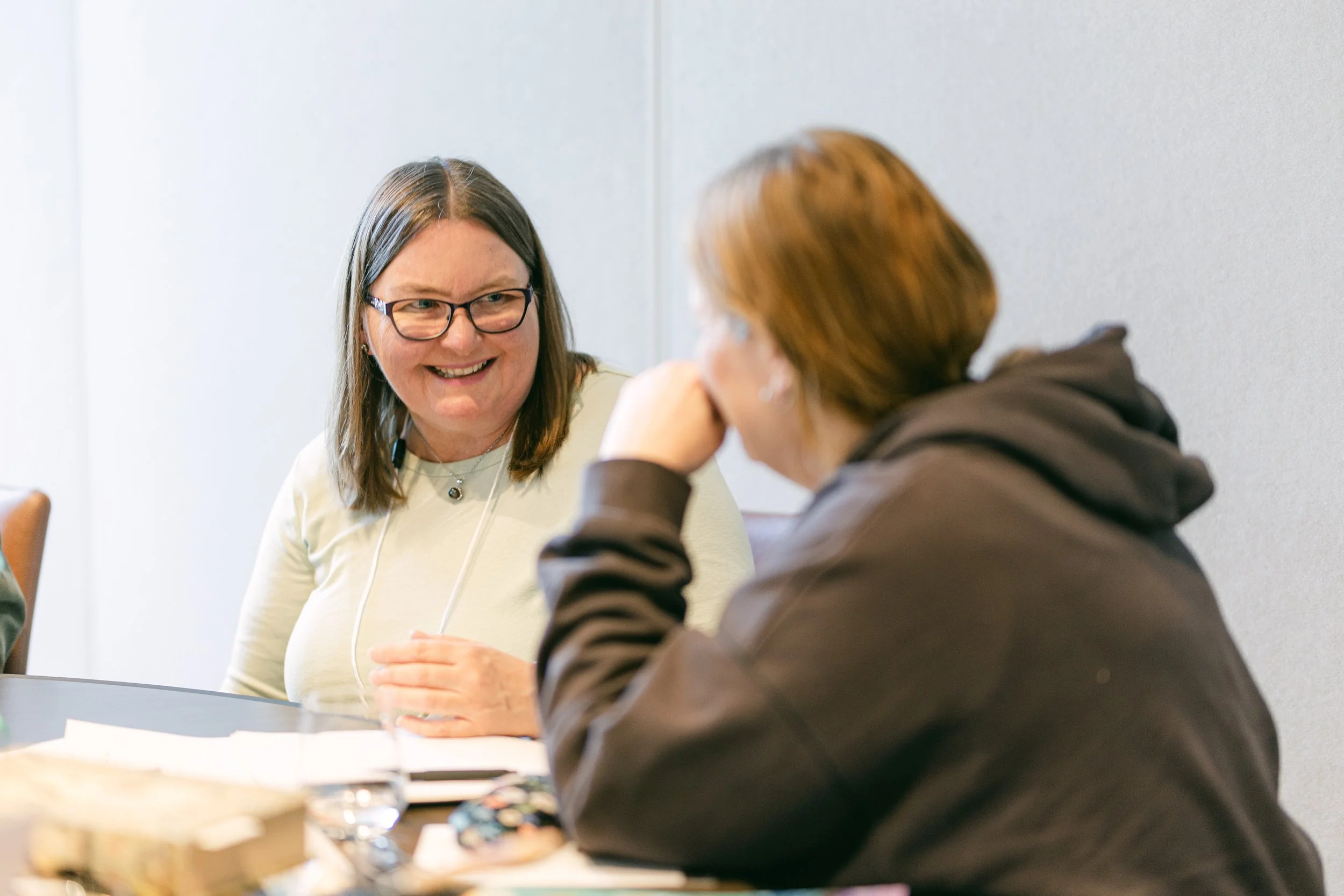 Two women sitting at a table engaged in a conversation, smiling, with papers and a glass of water in front of them.