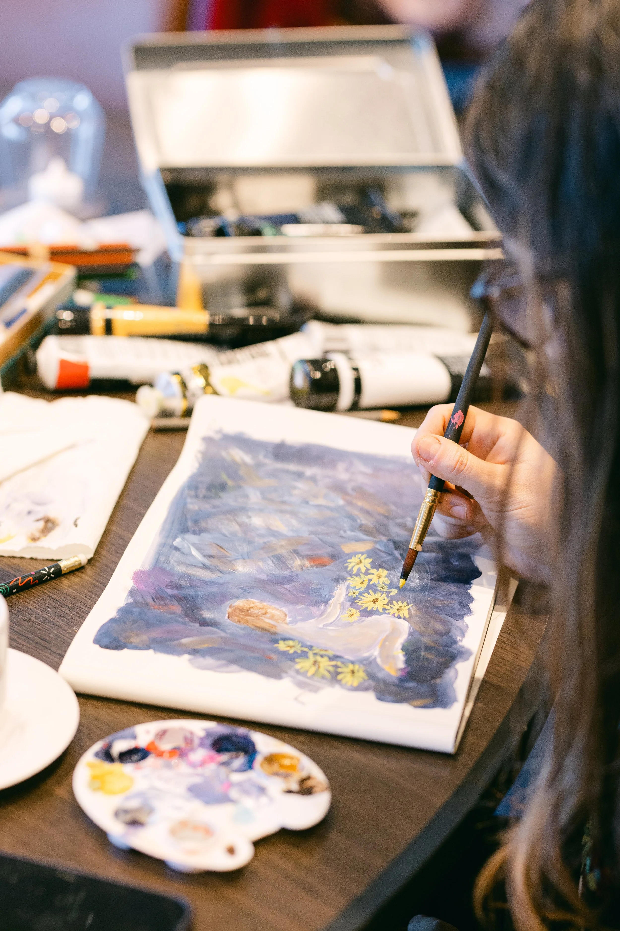 A person painting a landscape scene on paper with a paintbrush, surrounded by art supplies including paint tubes, a palette with mixed colors, and a silver box on a wooden table.