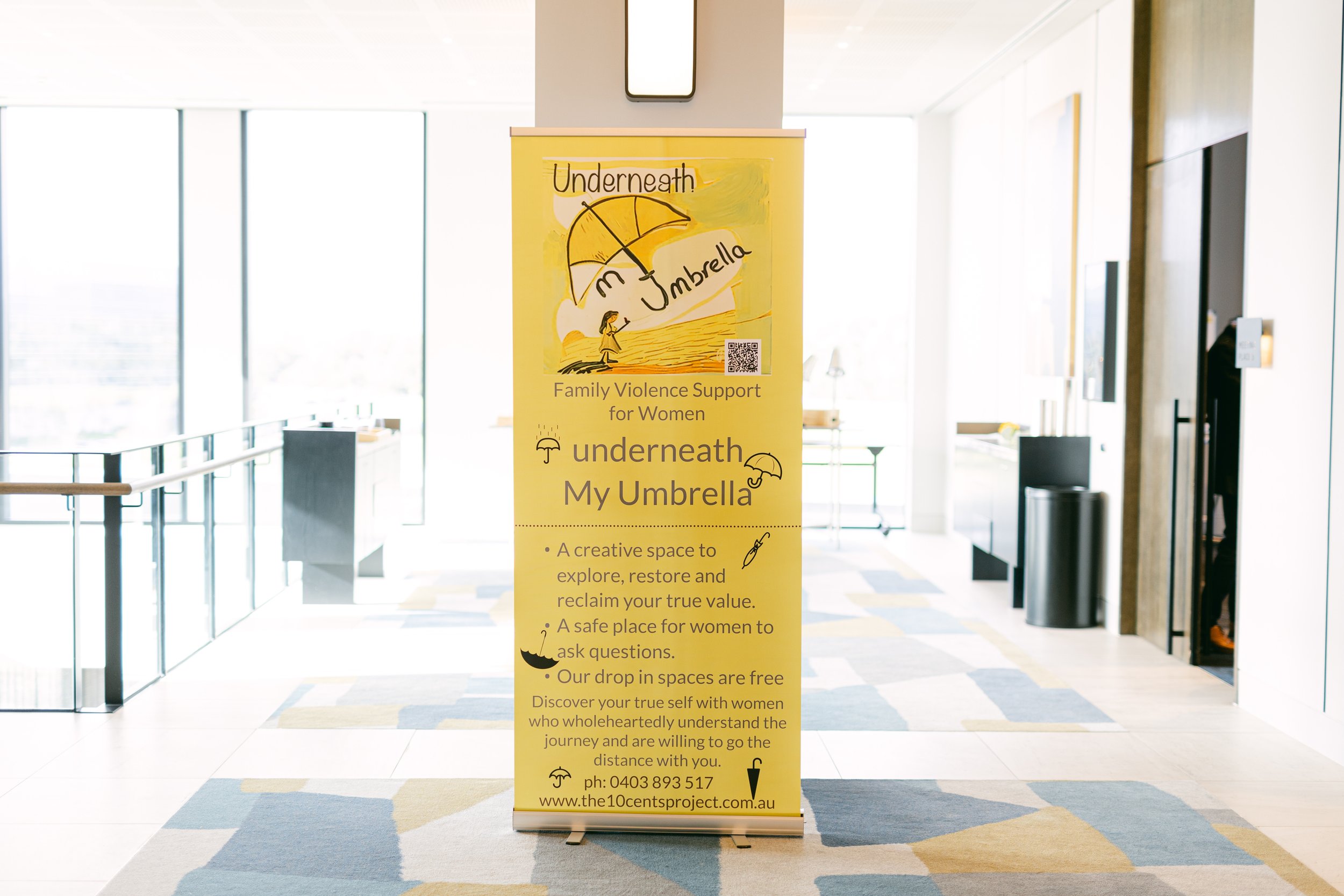 Yellow standing banner promoting family violence support for women called "Underneath My Umbrella," featuring illustrations of umbrellas, a woman, and a boat, with informational text about a safe space for women to explore and reclaim their value, in