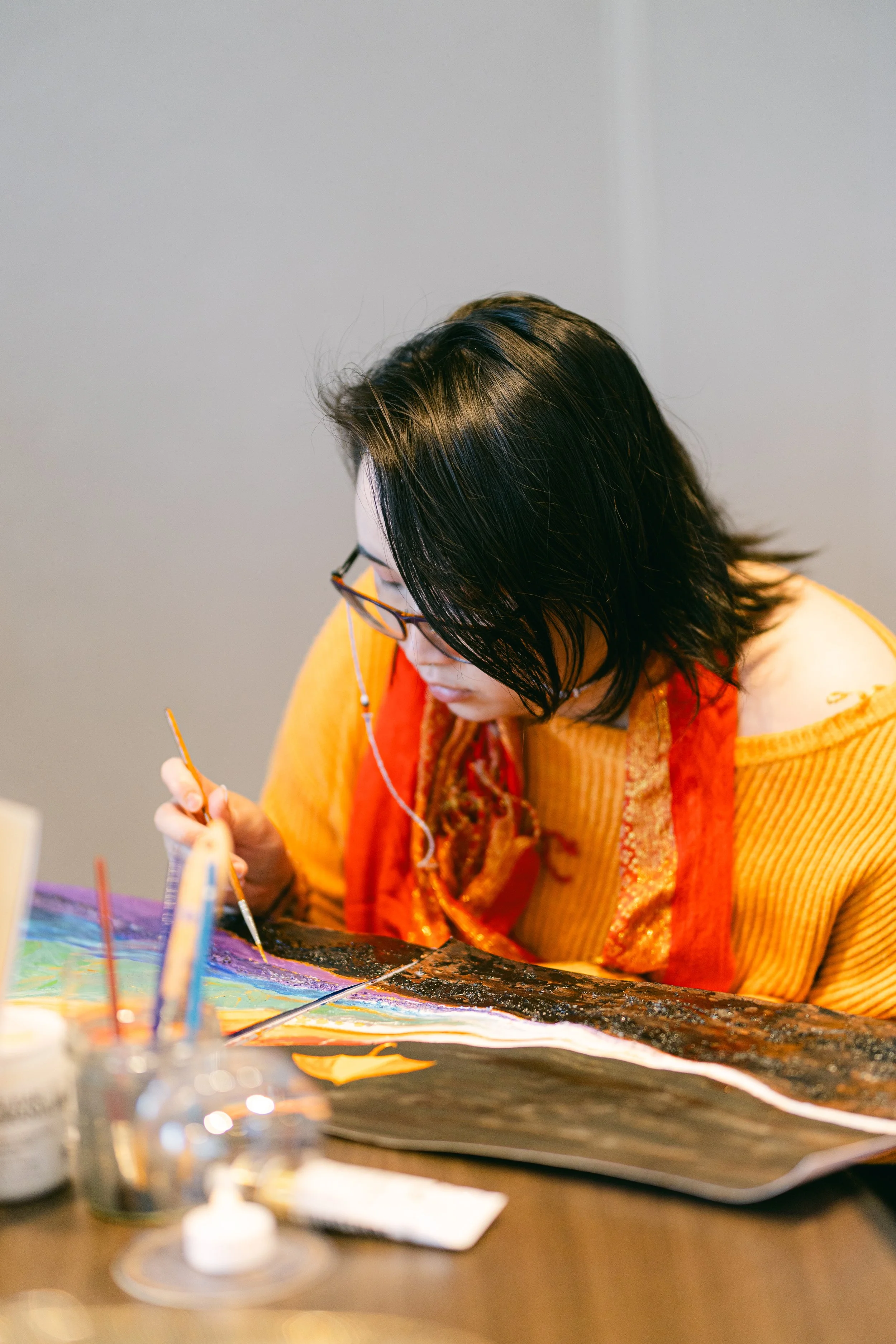 A woman with shoulder-length black hair, wearing glasses and an orange-yellow sweater with a red scarf, is painting with a small brush on a large canvas or paper on a table. Art supplies and a palette are visible in the foreground.