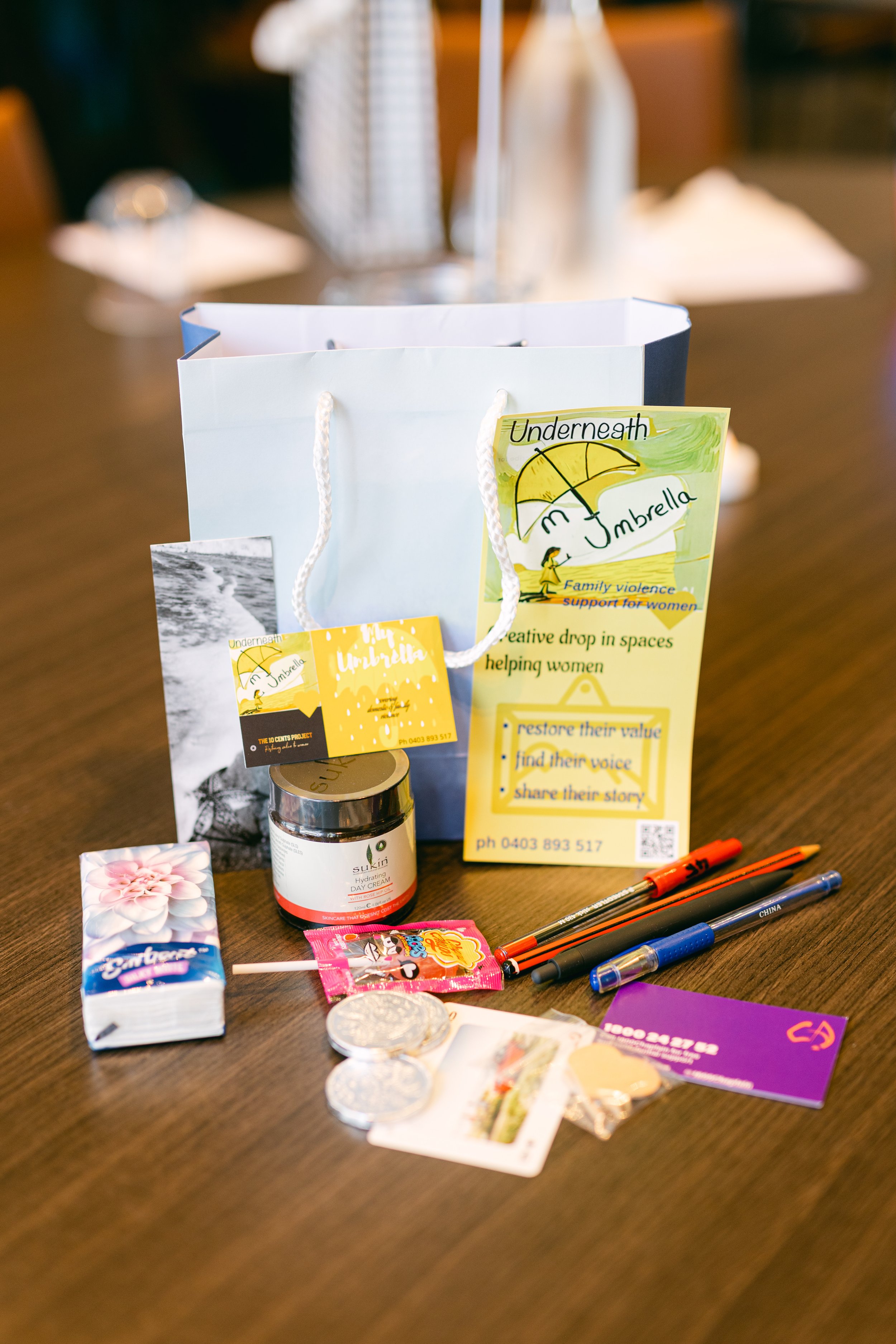 A gift bag on a table with promotional materials, a jar of cream, candies, and pens, likely for a women's support event.