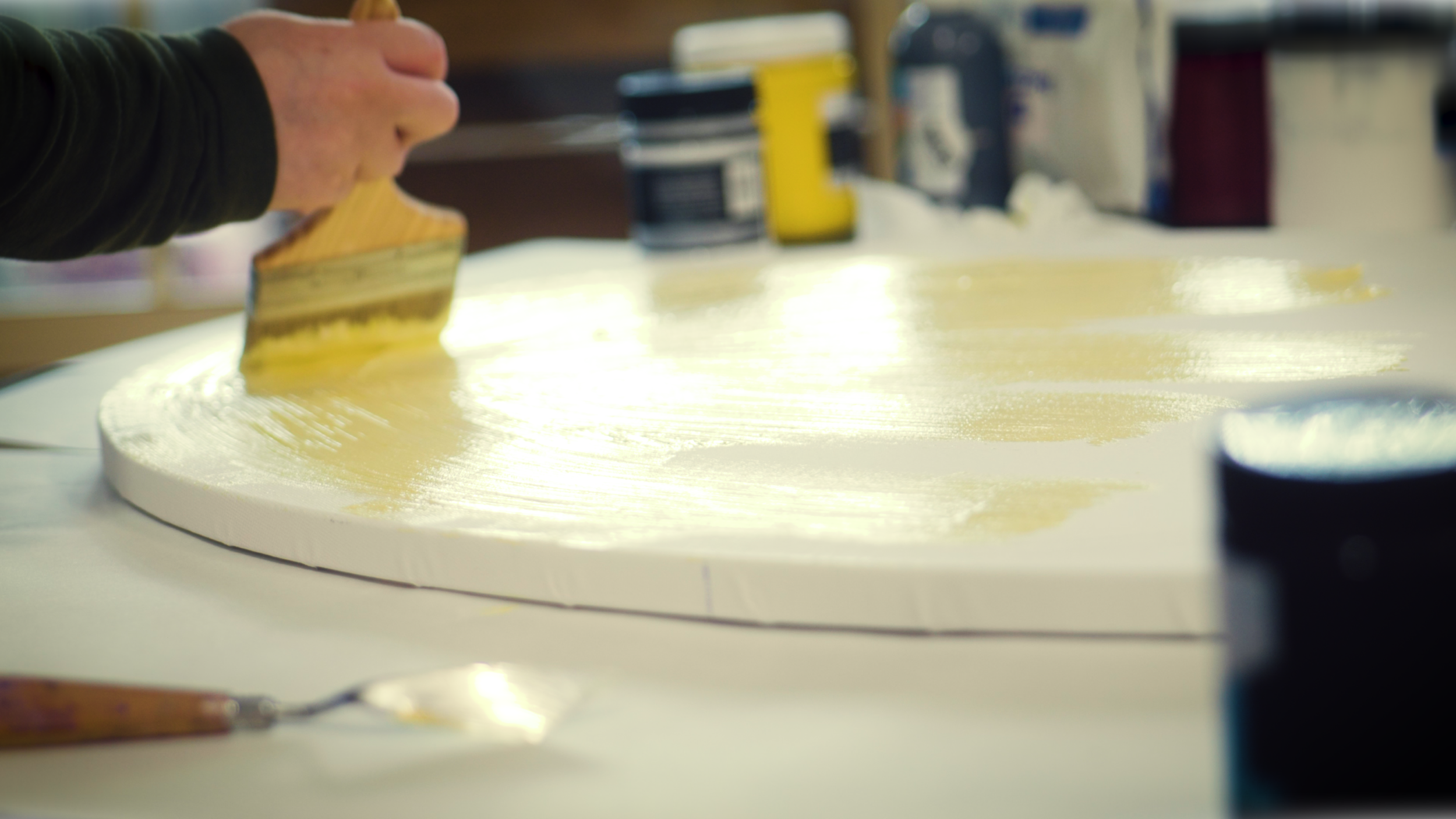 A person is spreading yellow paint on a white circular surface with a wide brush.