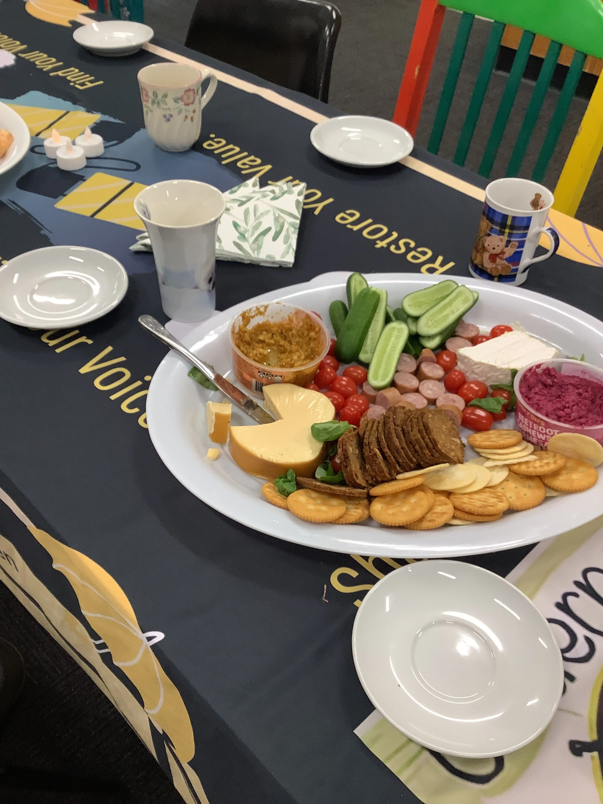 A platter with cucumber slices, cherry tomatoes, green bell pepper slices, a block of cheese, crackers, sausage slices, and dips on a table setting with small plates, cups, and a bib.