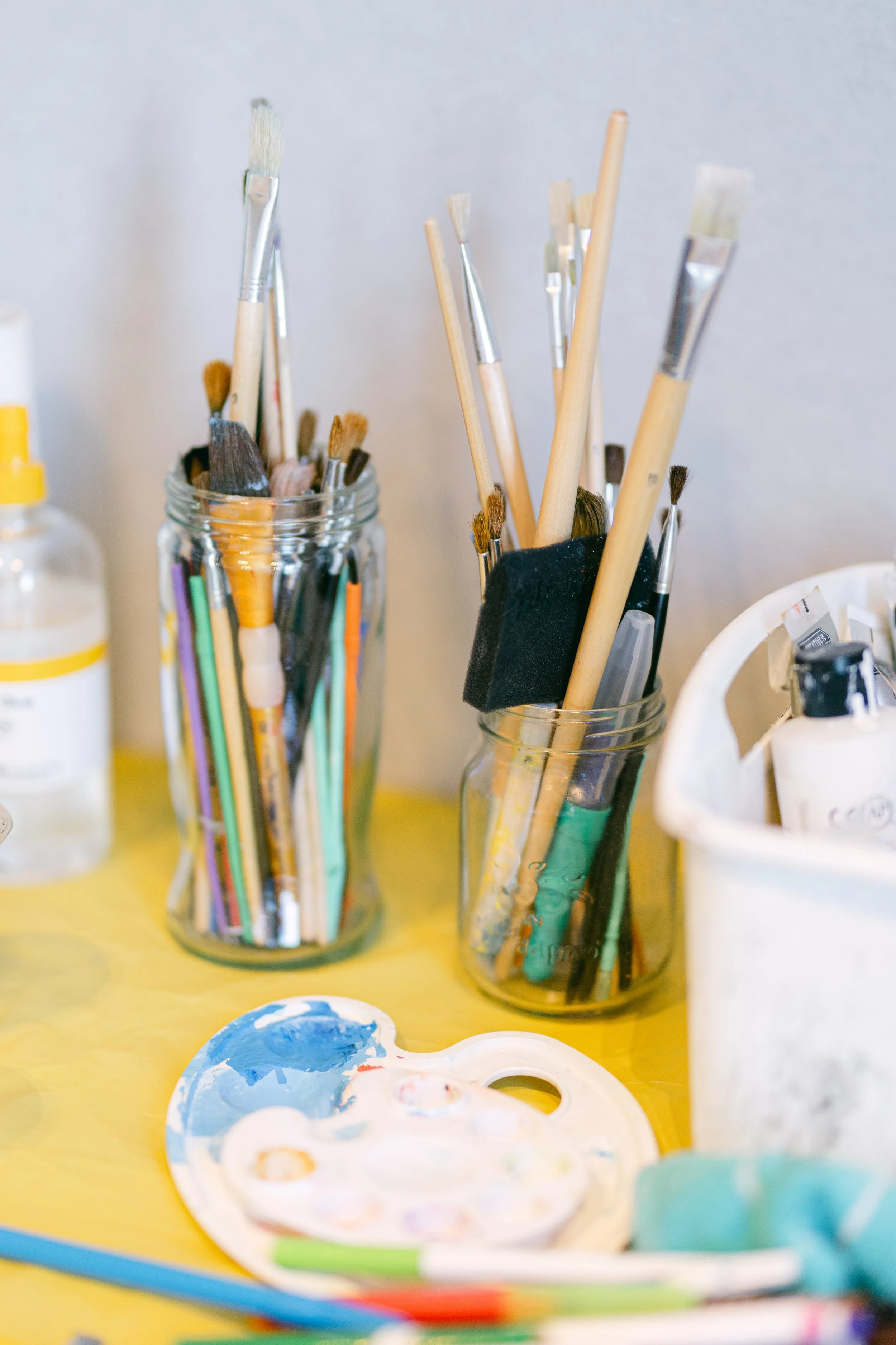 Paintbrushes and art supplies in glass jars placed on a yellow surface.