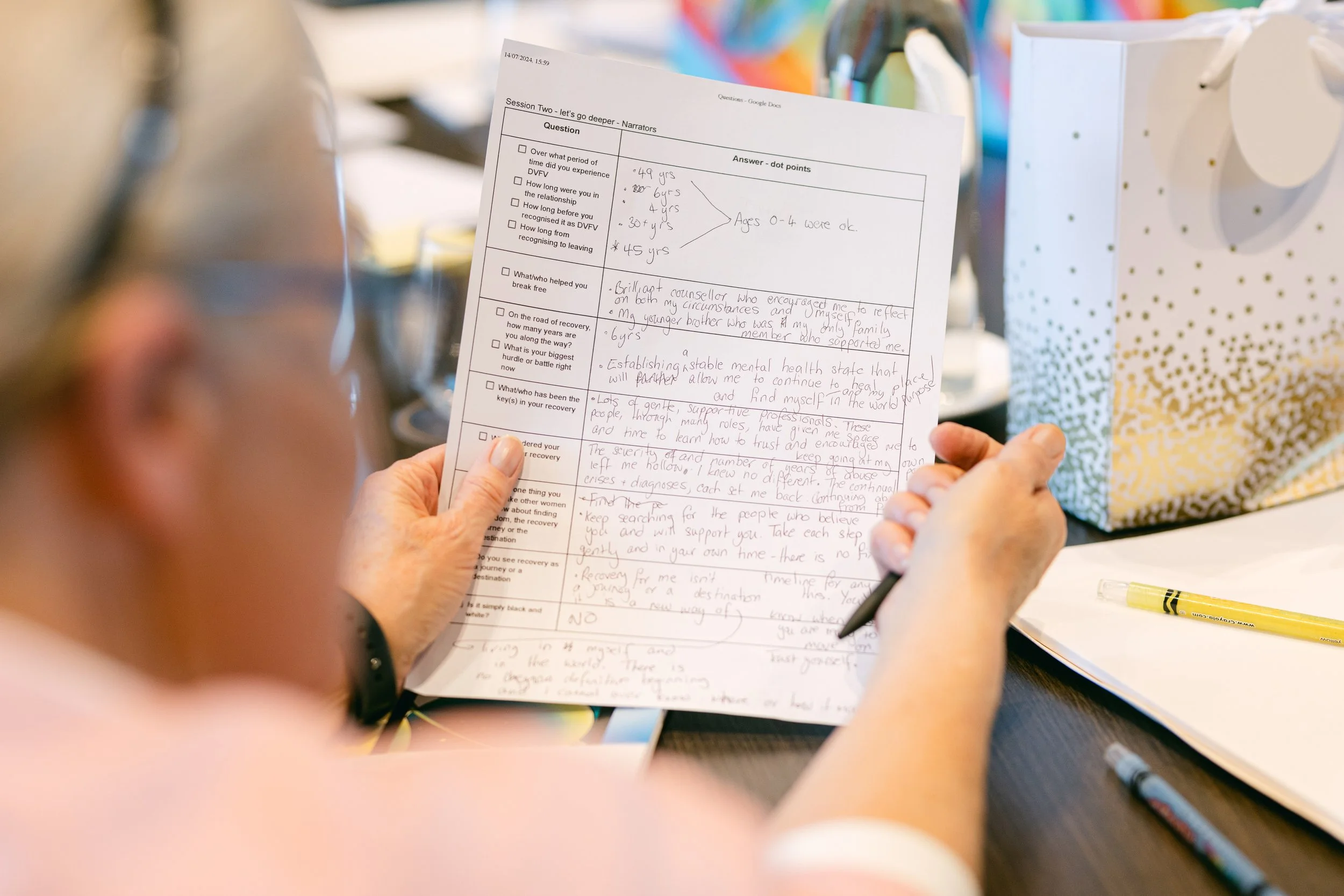 A person holding a sheet with handwritten notes and answers, sitting at a table with pens, a white notebook, and a decorative paper bag nearby.