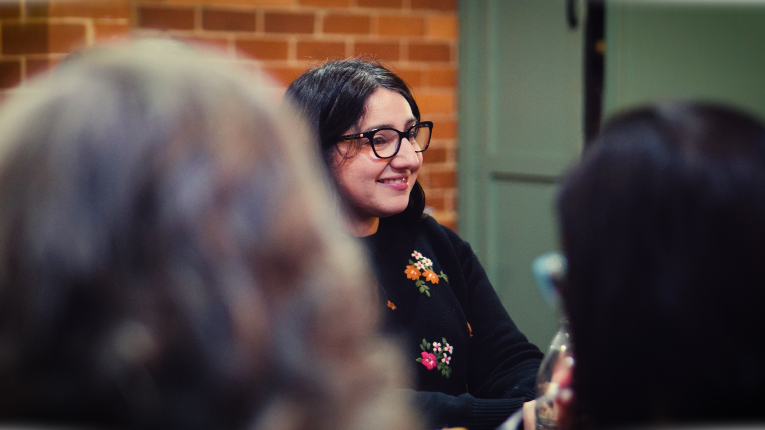 A woman with dark hair, glasses, and embroidered floral sweater smiling in a social setting with people in the foreground.