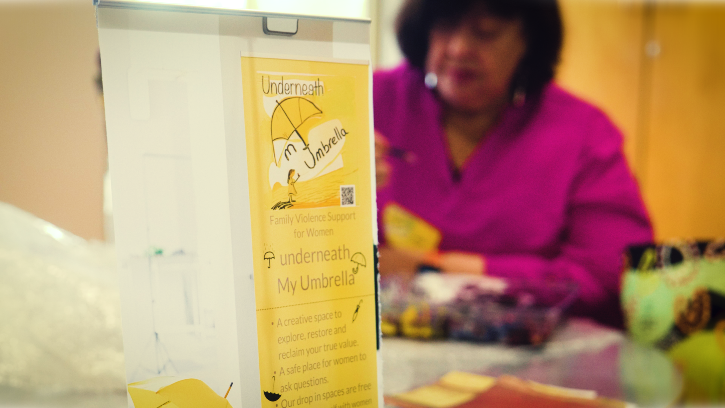 A woman in a pink top is seen at a table with colorful items, with a yellow informational display in the foreground promoting a family violence support project called 'Underneath My Umbrella'.