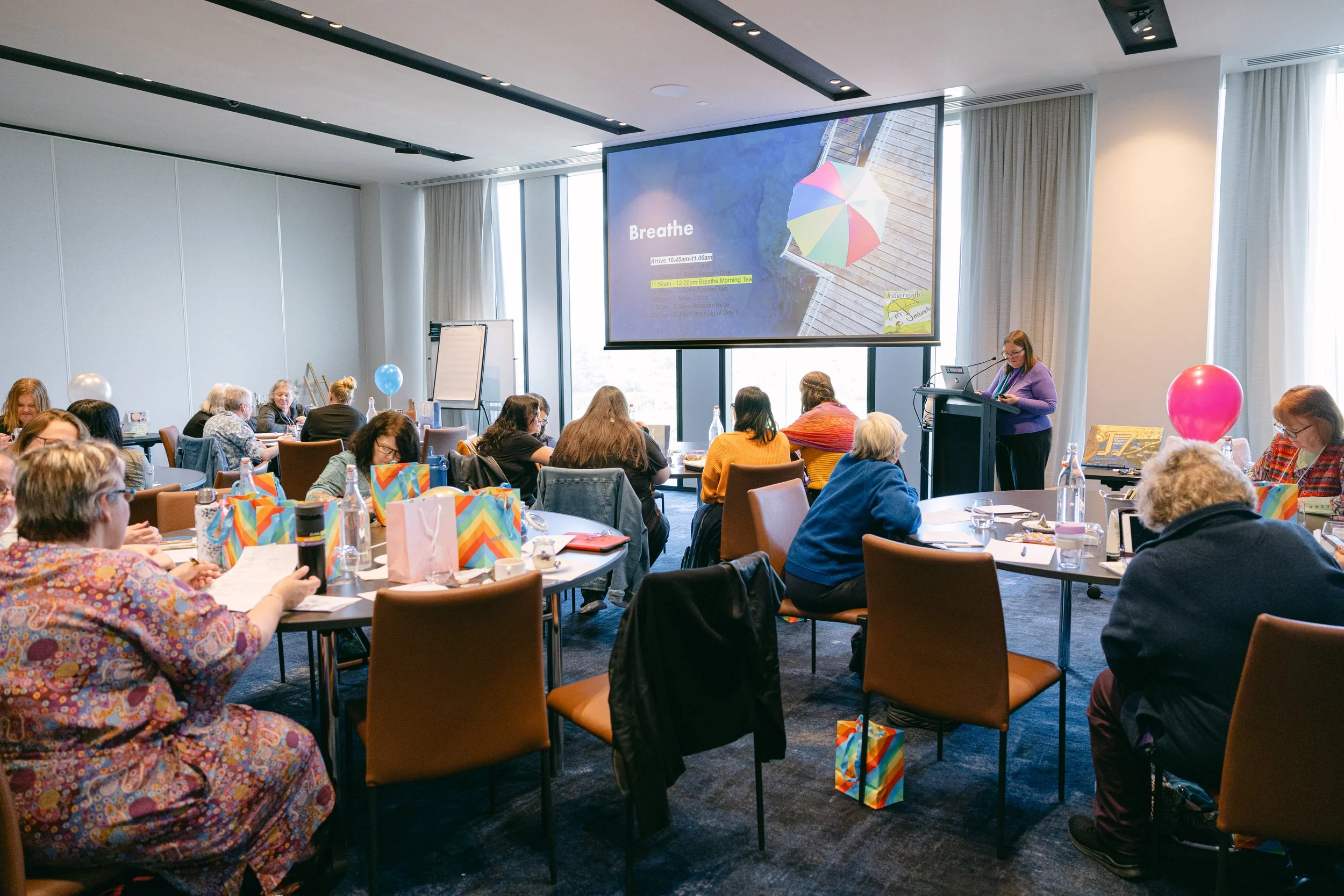 A conference room filled with people sitting at tables, listening to a speaker at a podium. The room is decorated with colorful gift bags and balloons. There is a large screen displaying a slide titled 'Breathe' with a photo of an umbrella on a deck.