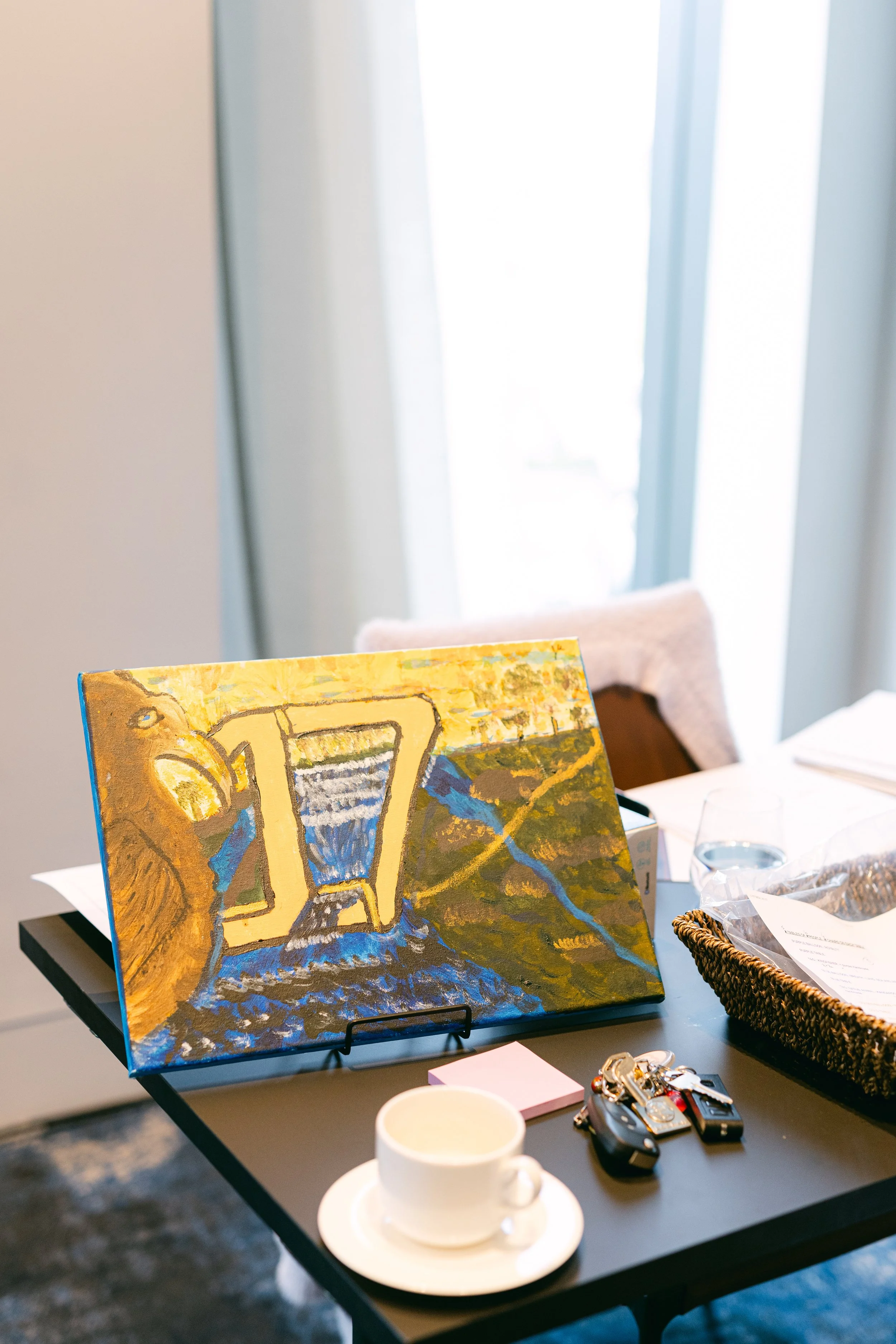 Painting of a waterfall and nature scene displayed on a table amidst various objects including keys, a cup, and papers.