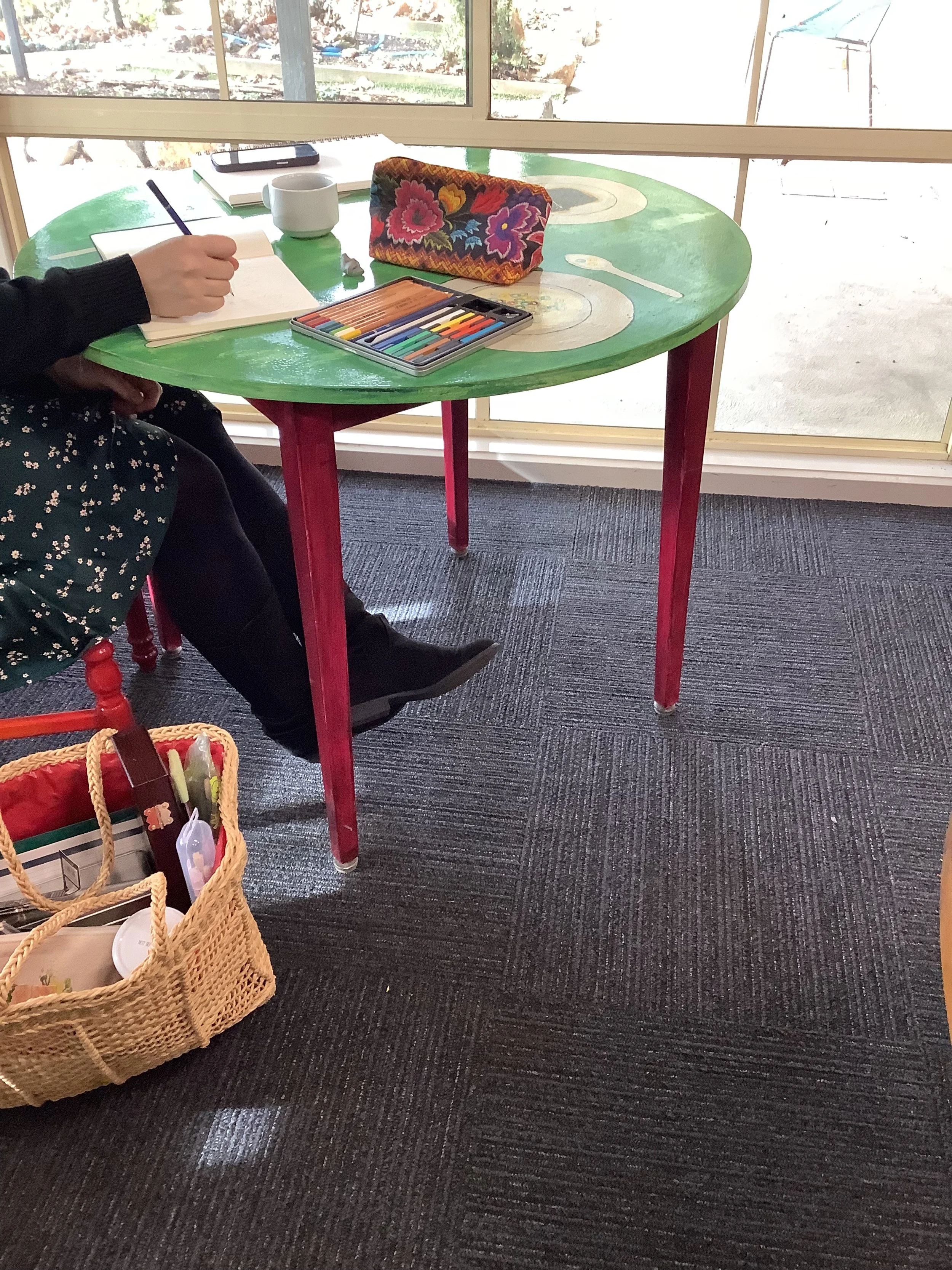 A round green table with red legs, set near a large window, holds a colorful floral pencil case, a set of colored markers, a notebook, a white mug, and a spoon, with a person partially visible writing in a notebook nearby.