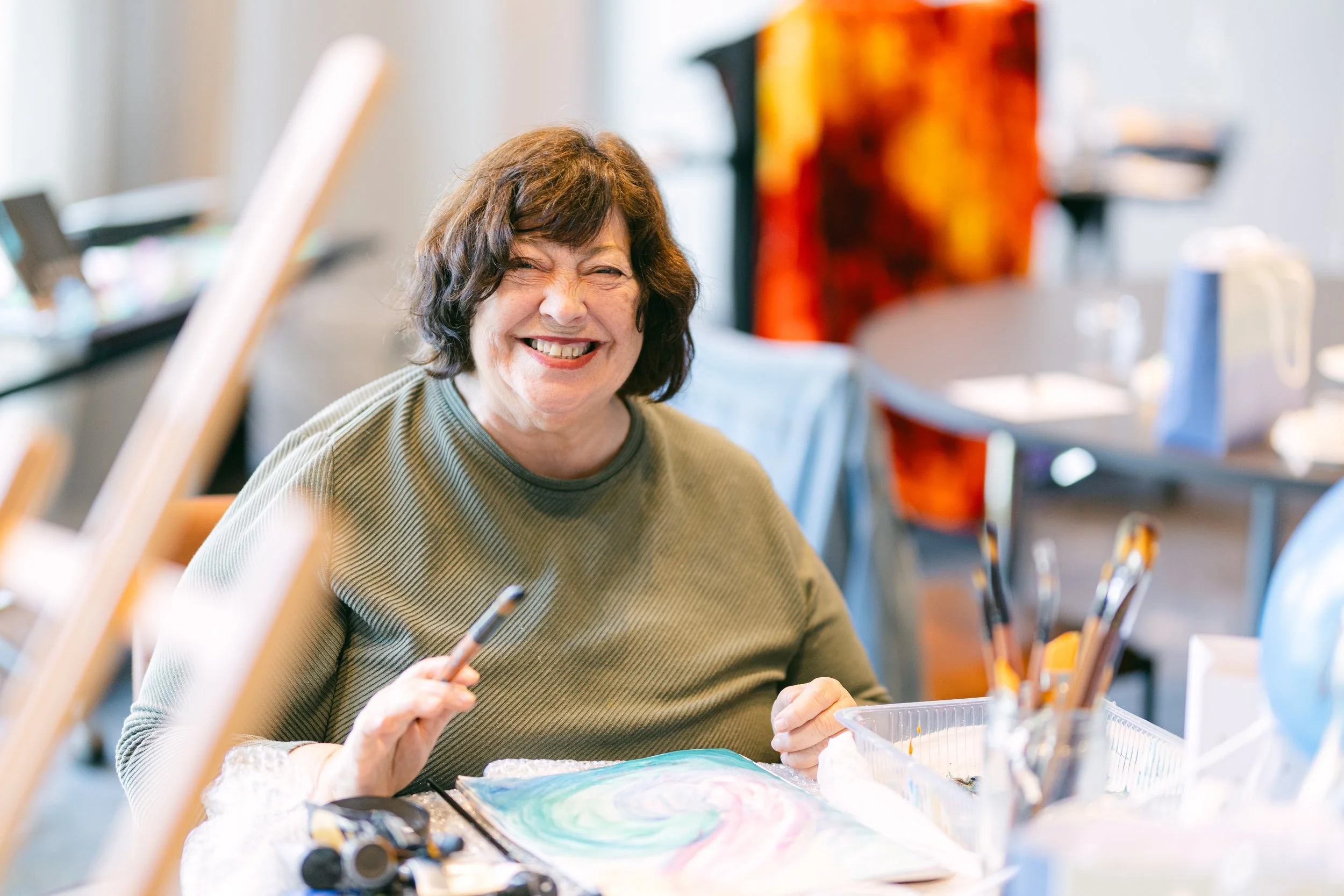 A woman with short dark hair smiling and laughing while sitting at a table with art supplies, including a colorful swirling painting, brushes, and a palette. The background shows a blurred indoor setting with art equipment and a brightly colored abst