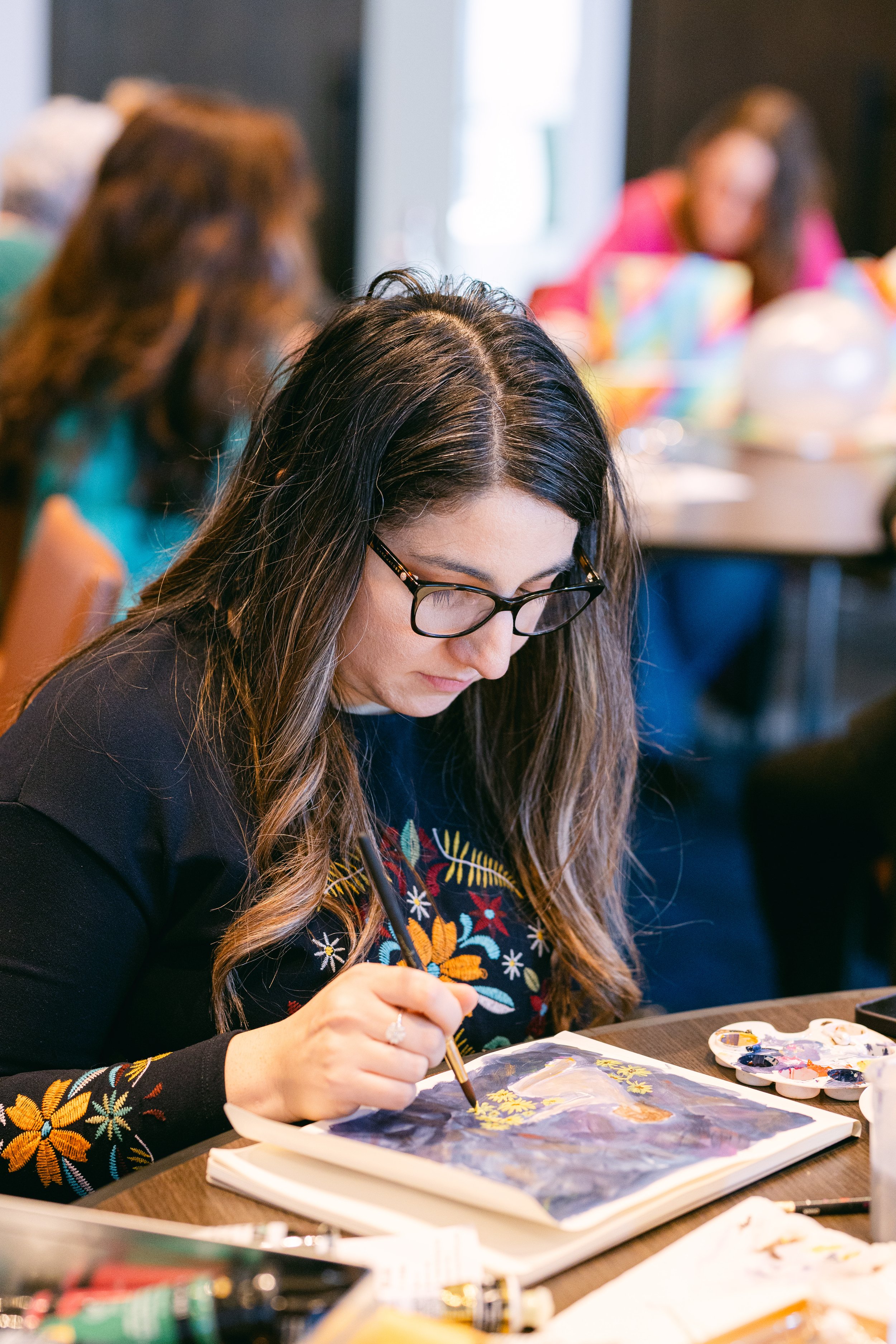 Woman painting in an art class, surrounded by other participants and art supplies.