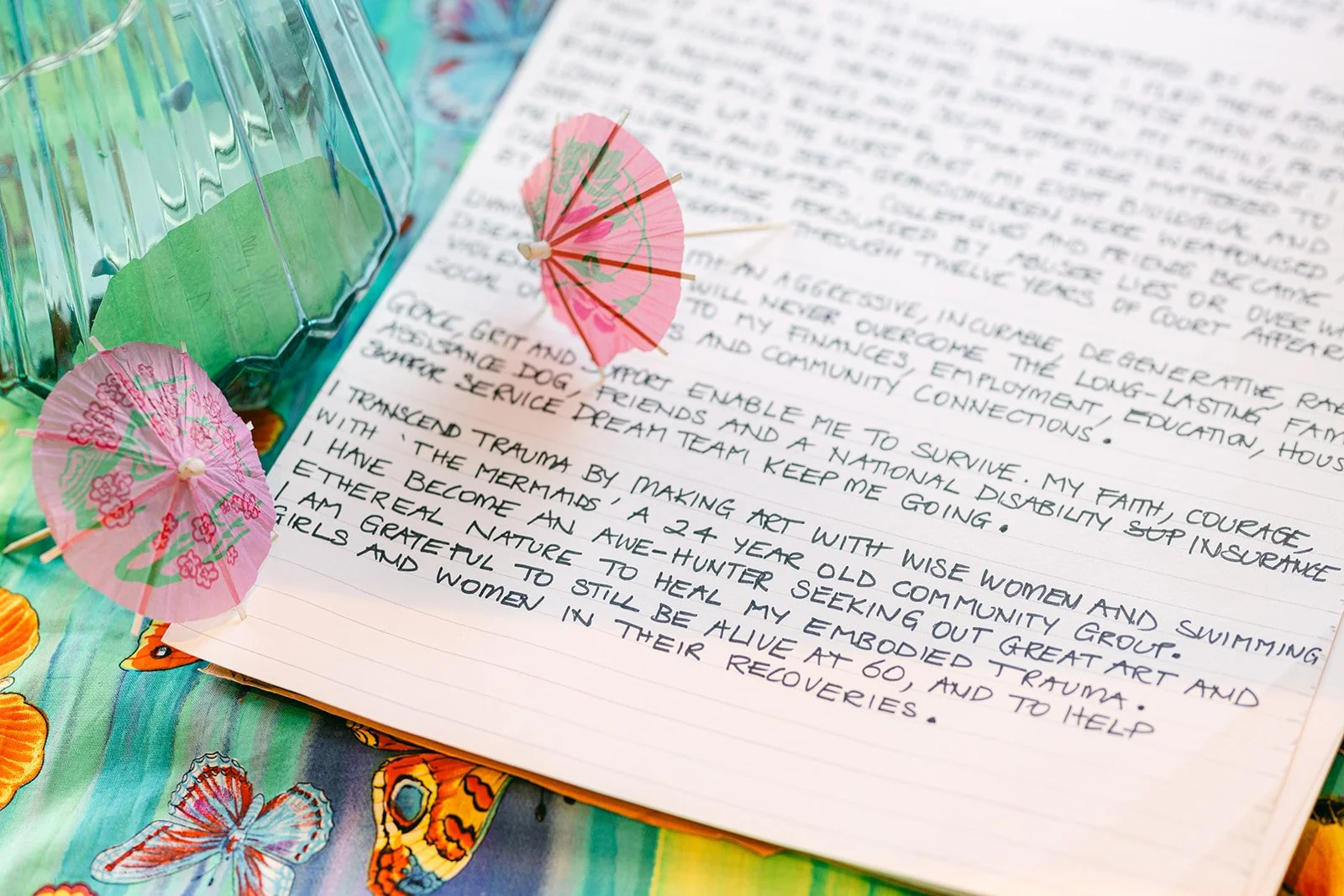 Close-up of handwritten letter on lined paper decorated with Asian-themed pink paper umbrellas and a green glass container, with a colorful butterfly-printed tablecloth underneath.