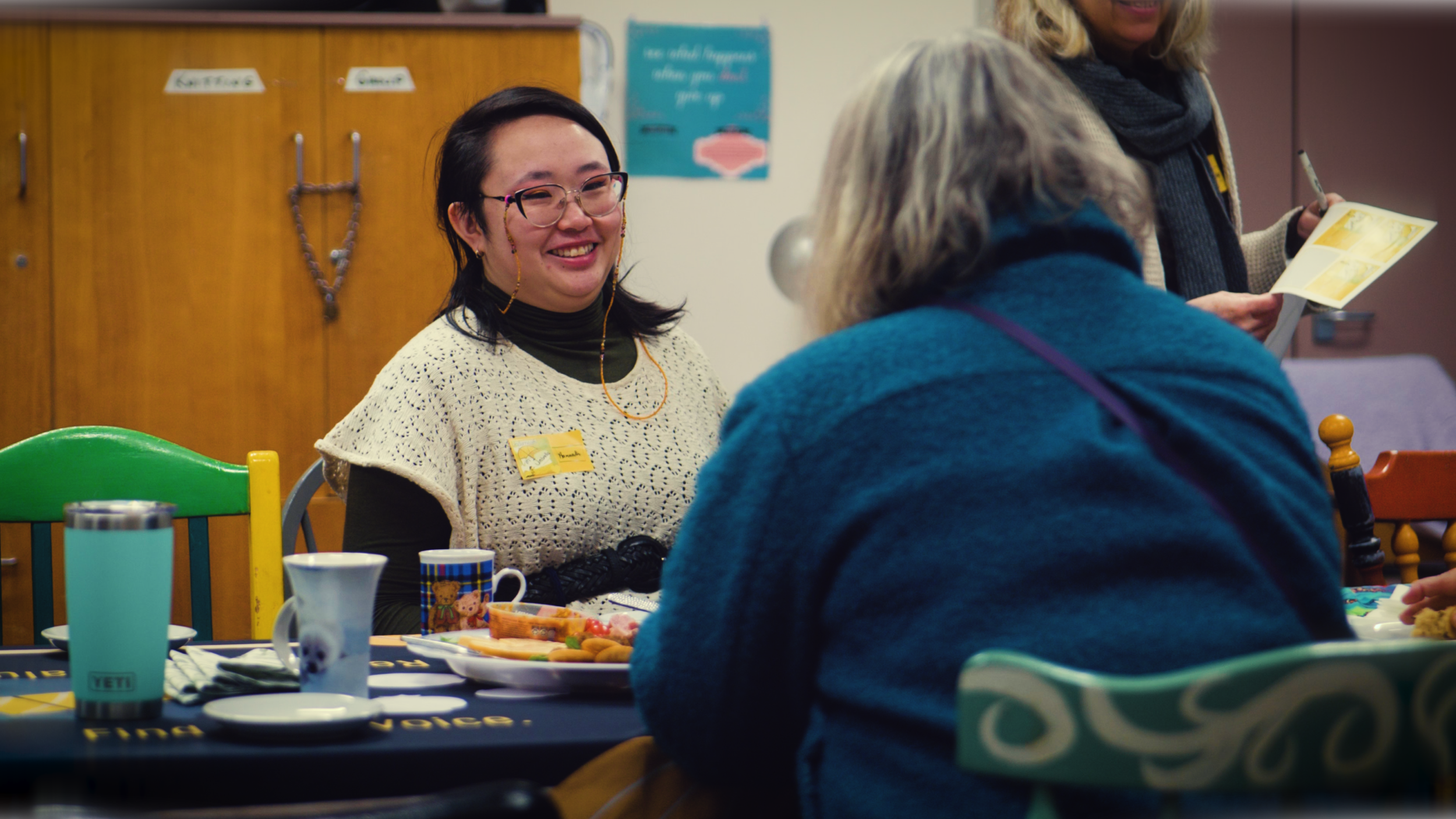 A woman with glasses and a white crochet top smiling at an elderly woman with gray hair, seated at a table with food and drinks, in a cozy indoor setting.