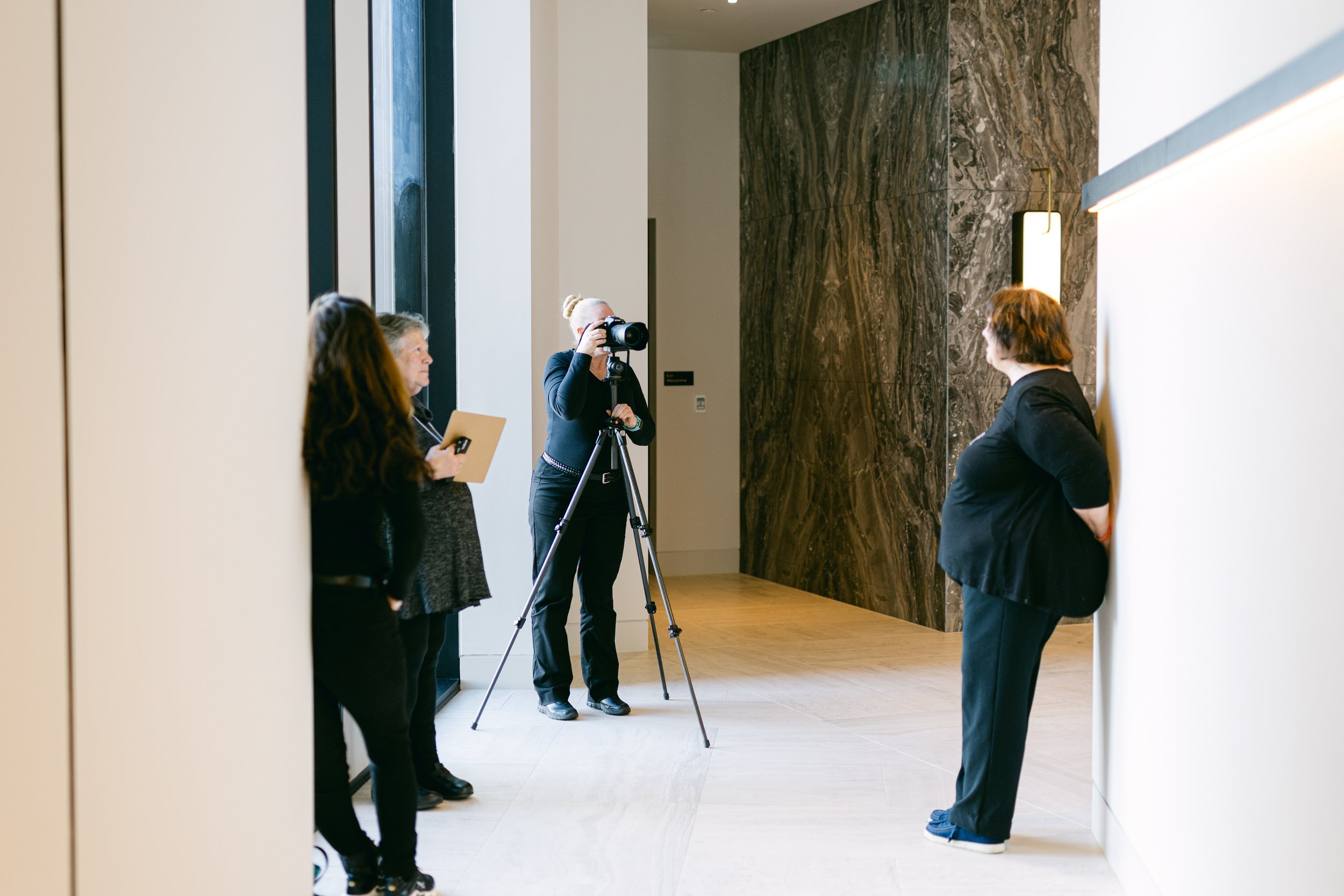 A woman in black with a camera on a tripod is taking a photo of a woman leaning against a wall, while three other women observe.
