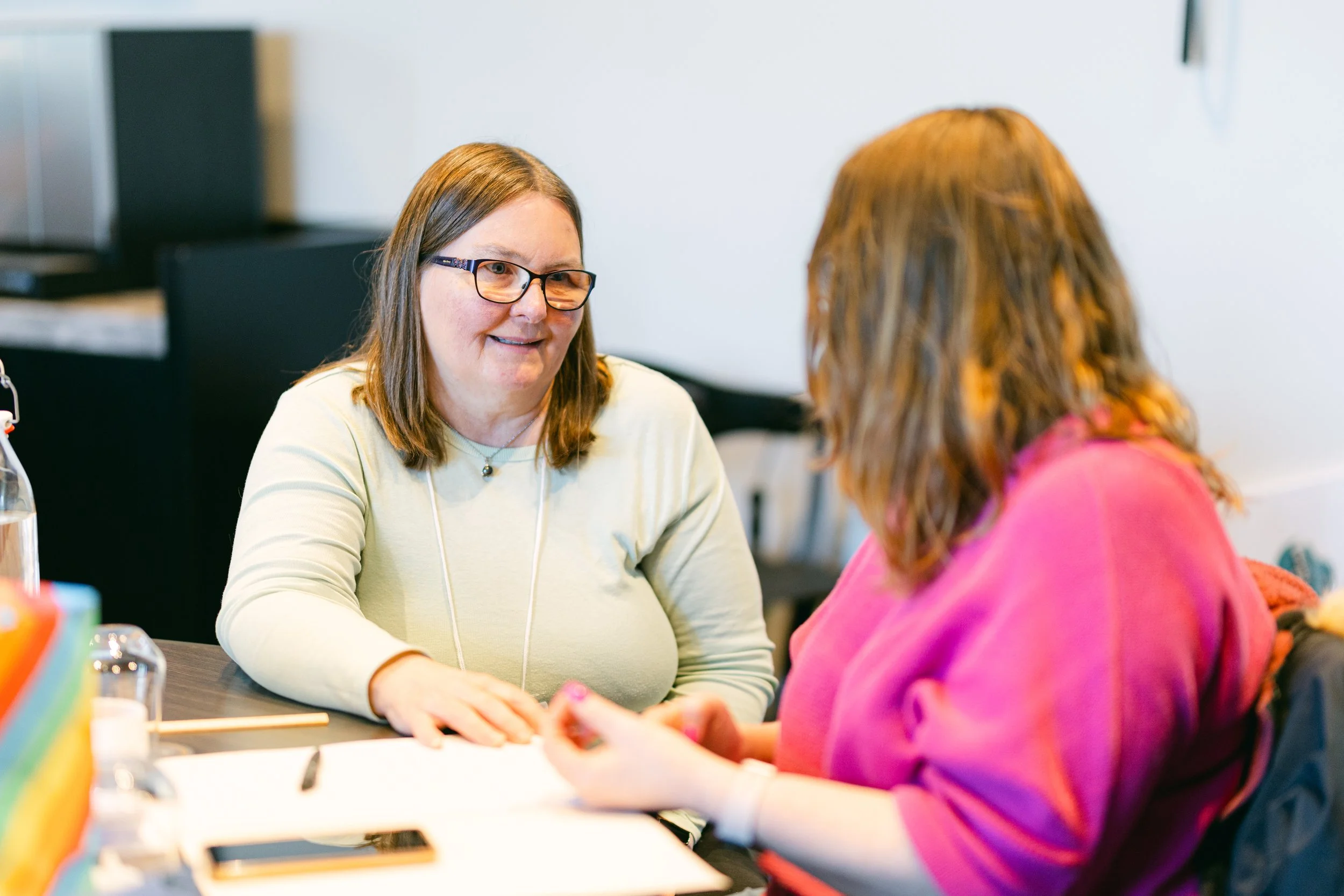 Two women engaged in conversation at a table, one gesturing with her hands, in a bright indoor setting.