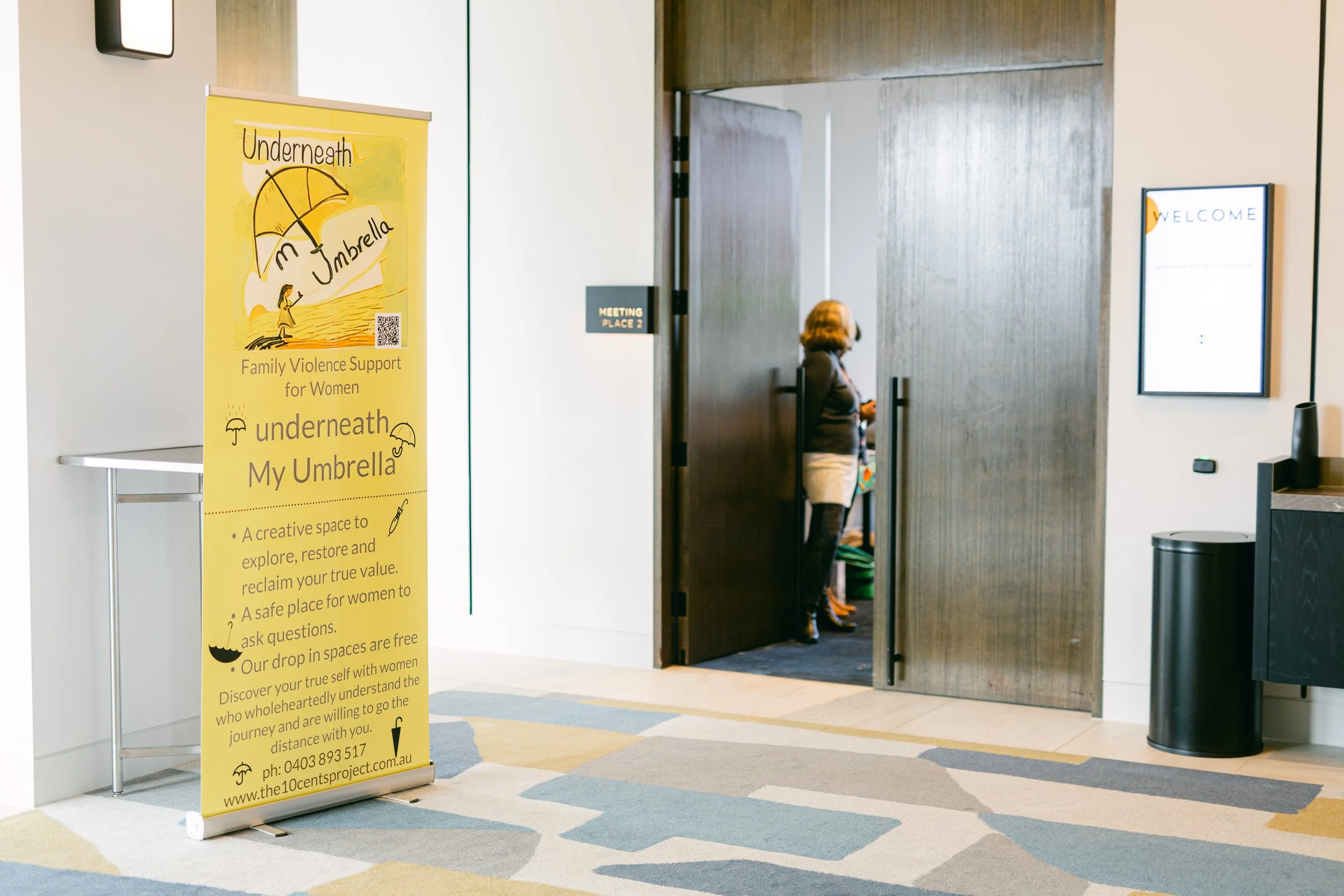 Yellow informational banner promoting 'underneath My Umbrella,' a space for women to explore, restore, and reclaim their value, located at a conference or event venue entrance. A woman stands partially visible inside a room with dark wooden door, nea