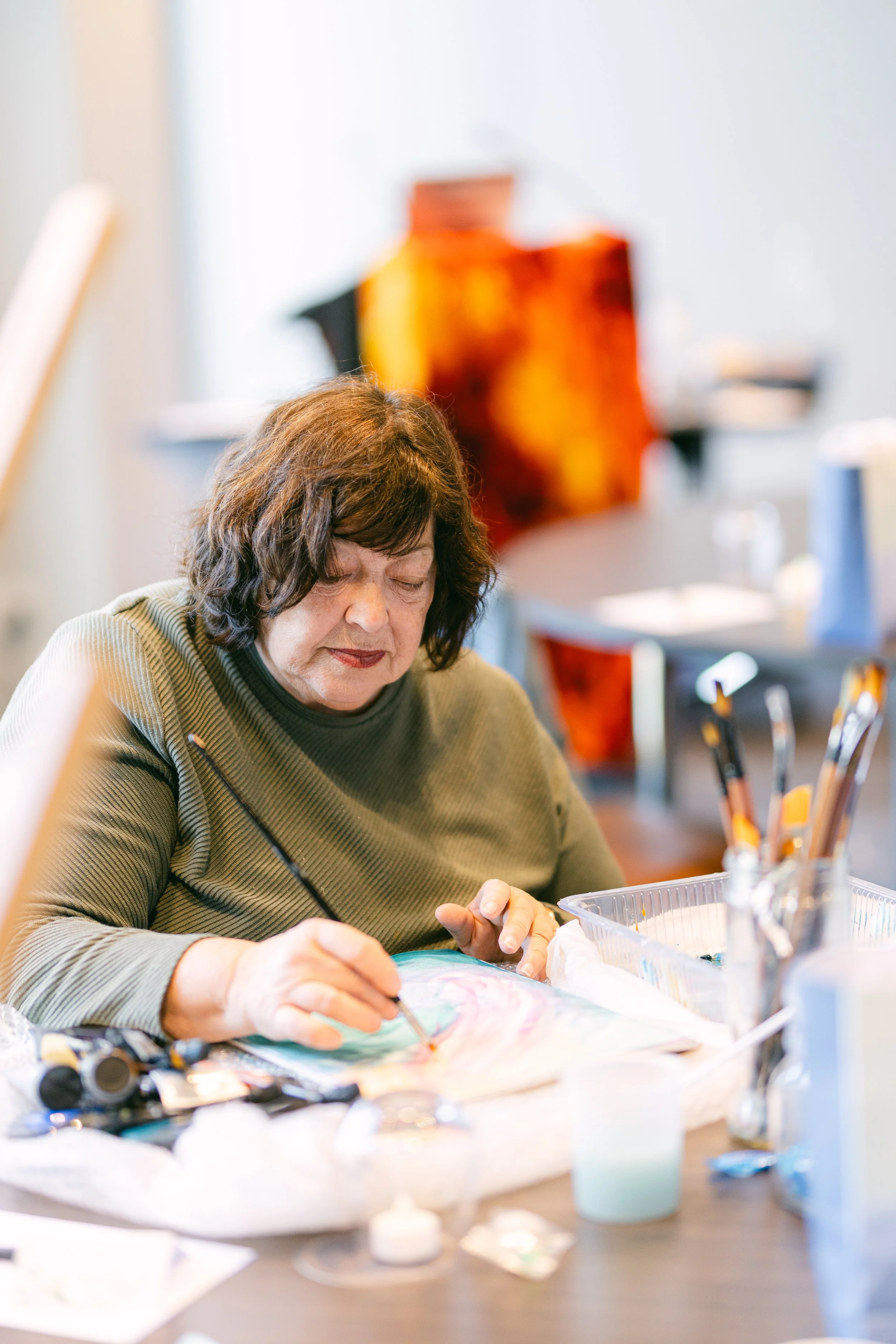 A woman painting with a paintbrush at a table filled with art supplies.