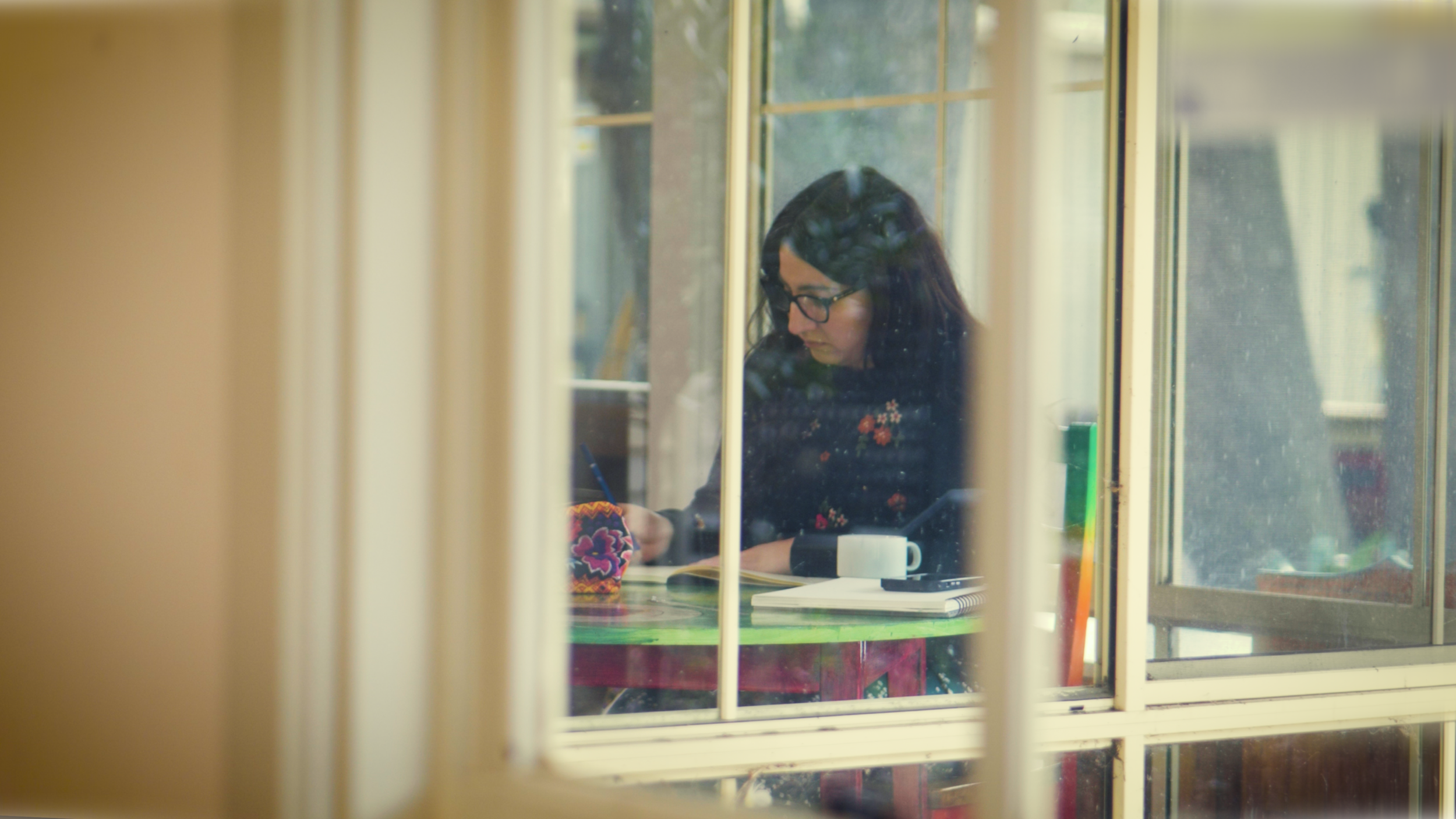 A woman with dark hair and glasses sitting at a table, seen through a window from outside, writing in a notebook with a coffee mug and papers on the table.